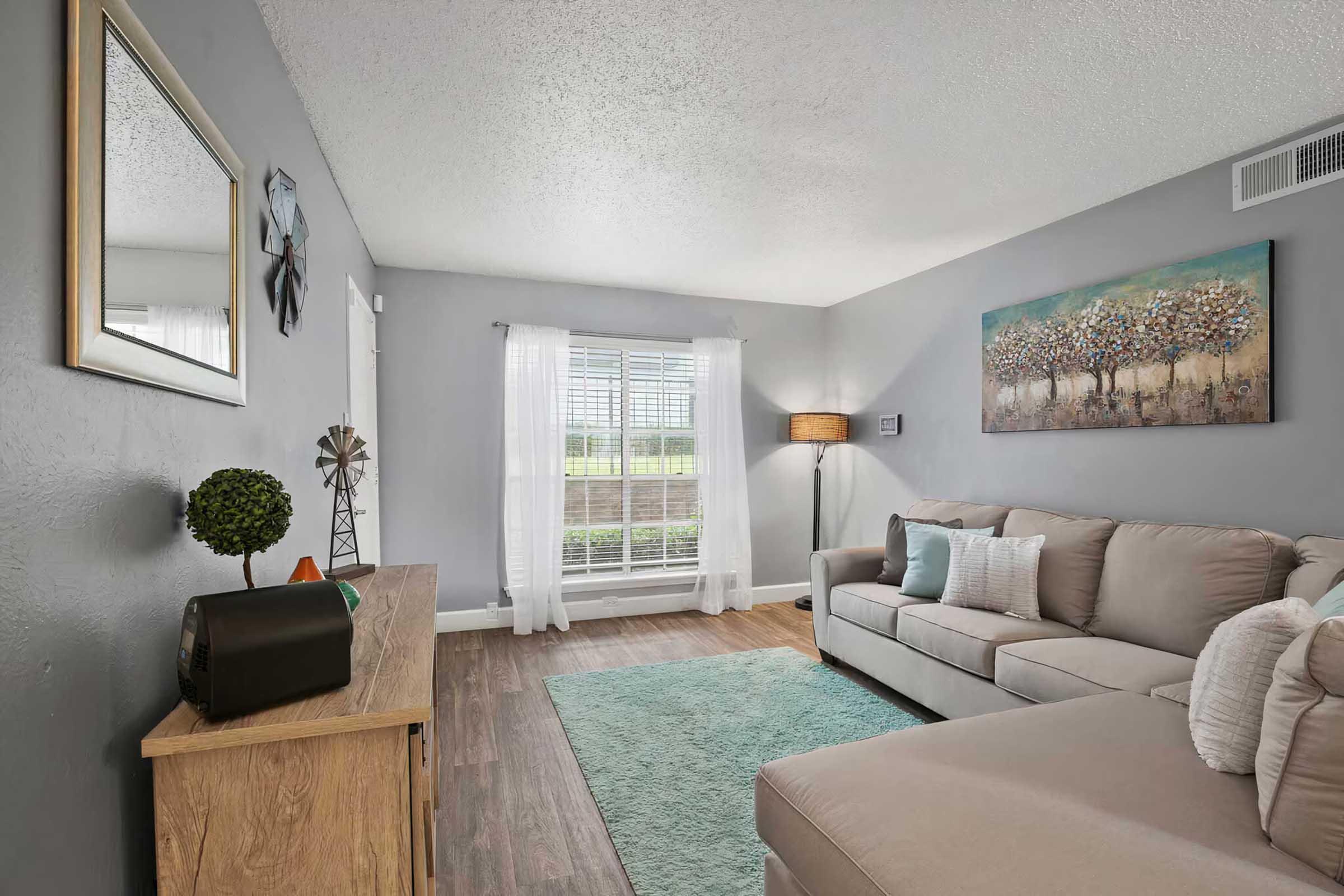 A cozy living room with light gray walls and wooden flooring. A large, comfortable beige sectional sofa is positioned in front of a window with sheer white curtains. A decorative painting of trees hangs on the wall, and a small green plant and a vintage radio sit on a wooden console table. Soft, teal area rug on the floor.