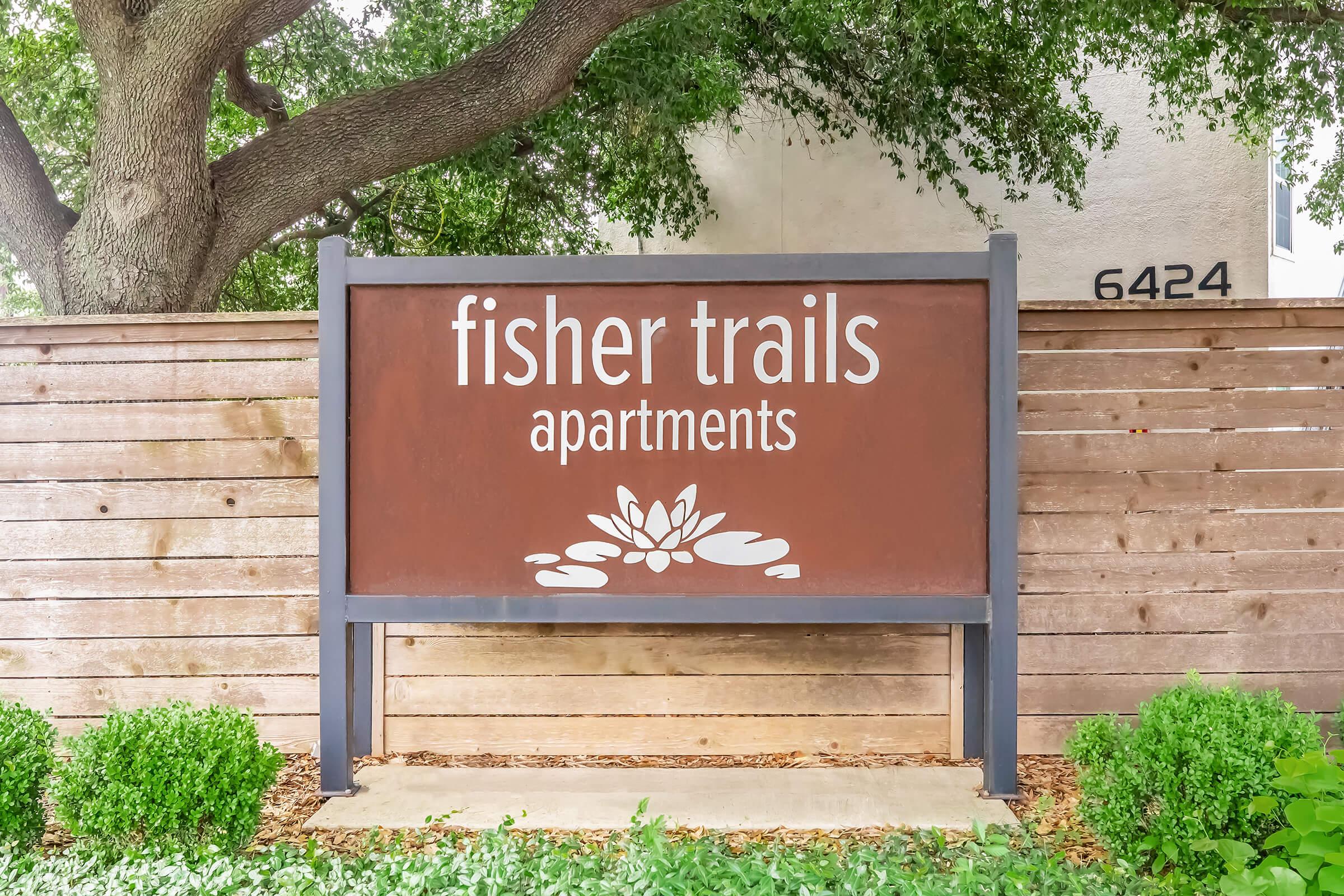 Sign for Fisher Trails Apartments, featuring a stylized lotus flower design. The sign is mounted on a wooden fence and includes the address "6424." Lush greenery surrounds the base, creating an inviting atmosphere.