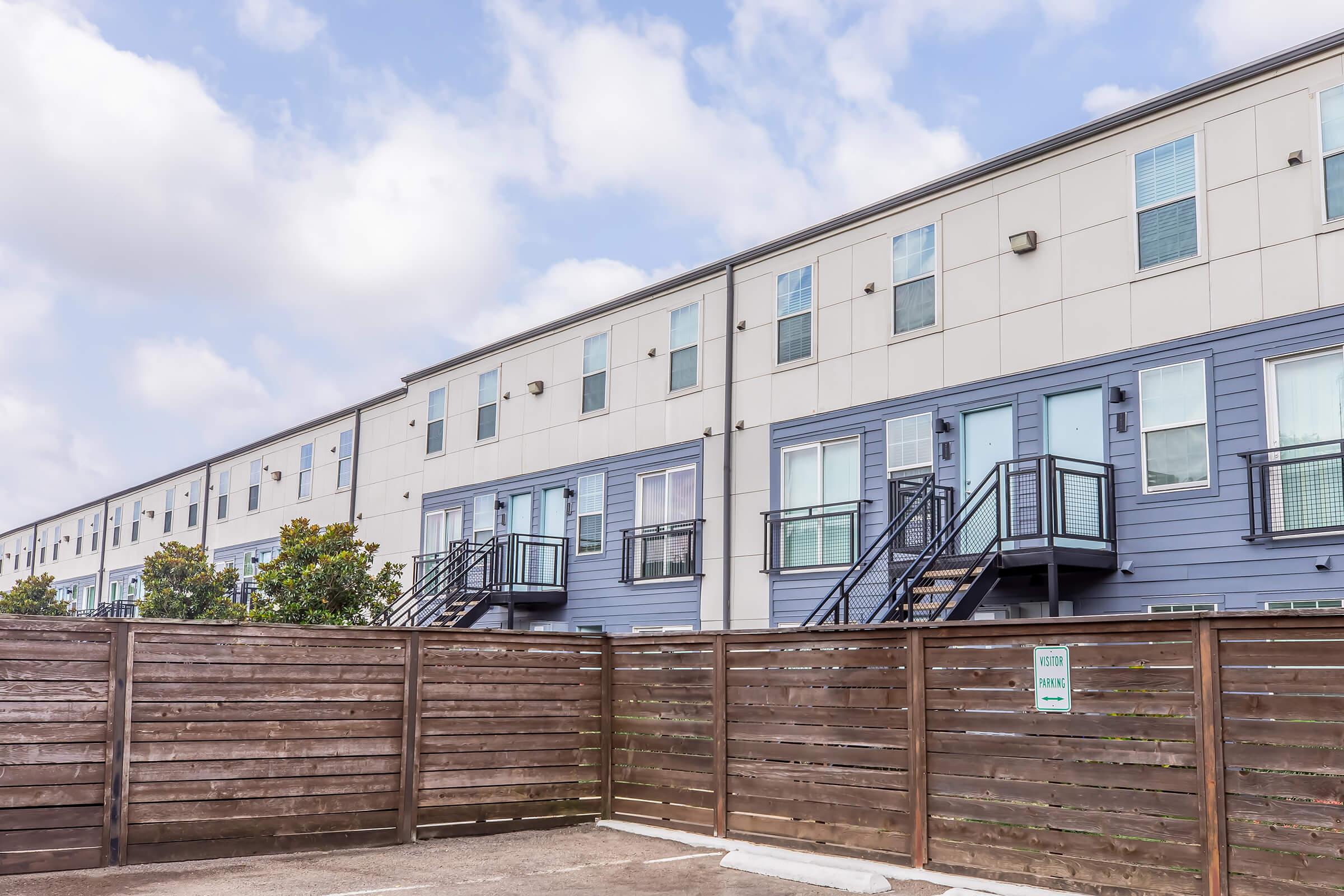 Row of modern, multi-unit residential buildings with blue and gray exteriors. Each unit features a private entrance with stairs leading up to a deck or balcony. A wooden fence with a sign is visible in the foreground, providing a sense of privacy to the area. The sky is partly cloudy.