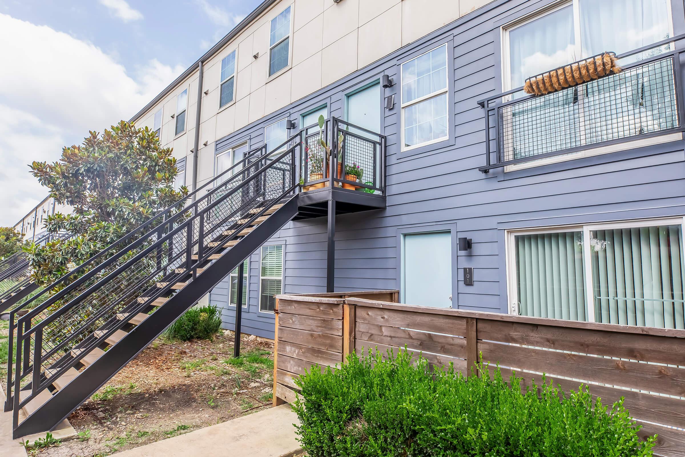 Exterior view of a modern apartment building featuring a staircase leading to upper floor units. The building has blue siding and large windows with plants on the balcony. A wooden fence and landscaped greenery are visible in the foreground. The sky is partly cloudy.