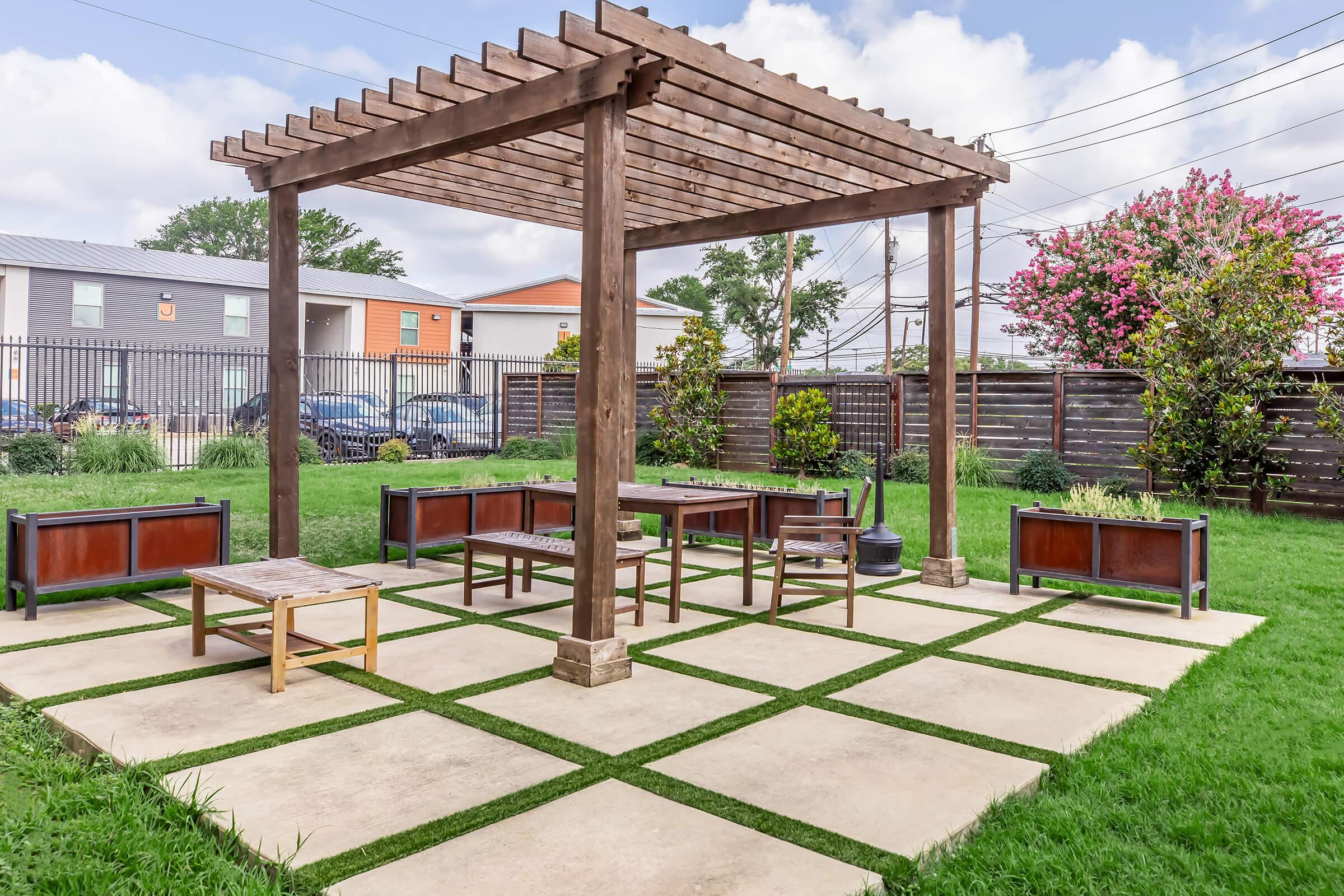 A landscaped outdoor area featuring a wooden pergola with a seating arrangement beneath it. The ground is paved with large stone slabs interspersed with grass. Flowering plants and shrubs are positioned around the perimeter, and a fence is visible in the background, alongside parked cars.