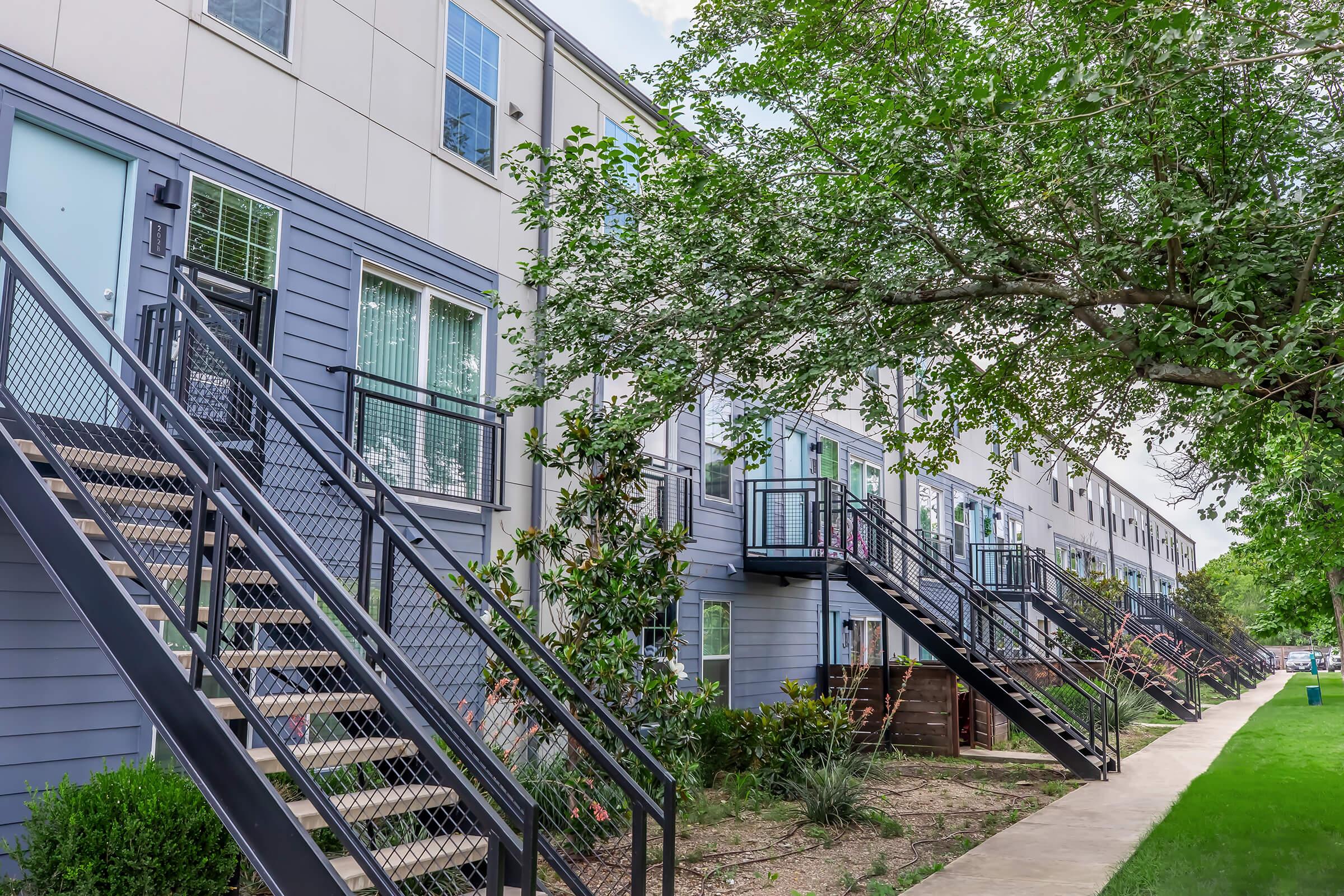 A row of modern multi-unit buildings with blue-gray siding and large windows. Each unit has a set of stairs leading up to a deck, surrounded by greenery and trees. The scene is set in a landscaped area, with a pathway running alongside the buildings.
