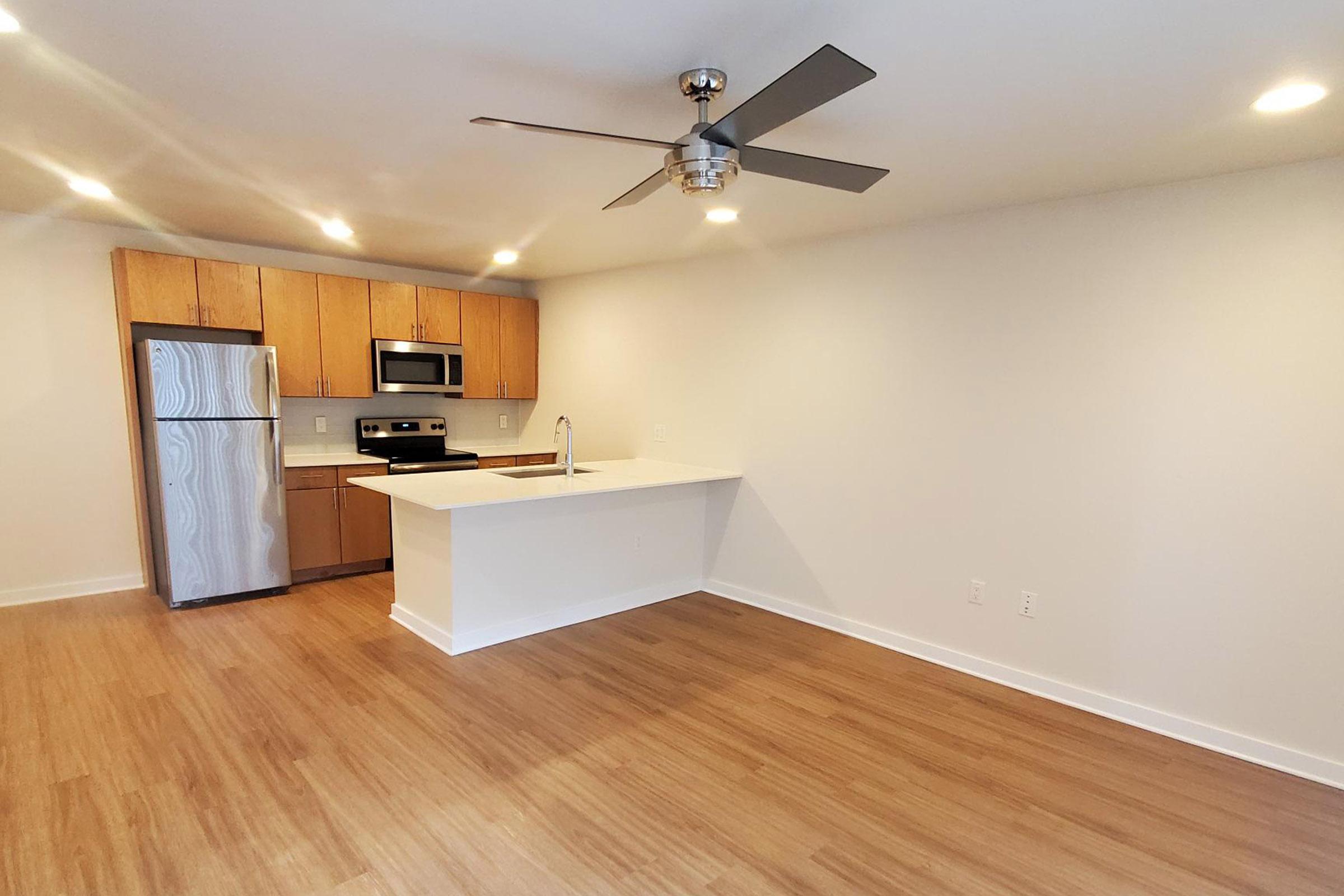 A bright, modern kitchen and living area featuring wood cabinetry, stainless steel appliances, and a spacious countertop. The ceiling has a contemporary ceiling fan, and the flooring is light wood. The walls are painted in a neutral shade, enhancing the open and airy feel of the space.