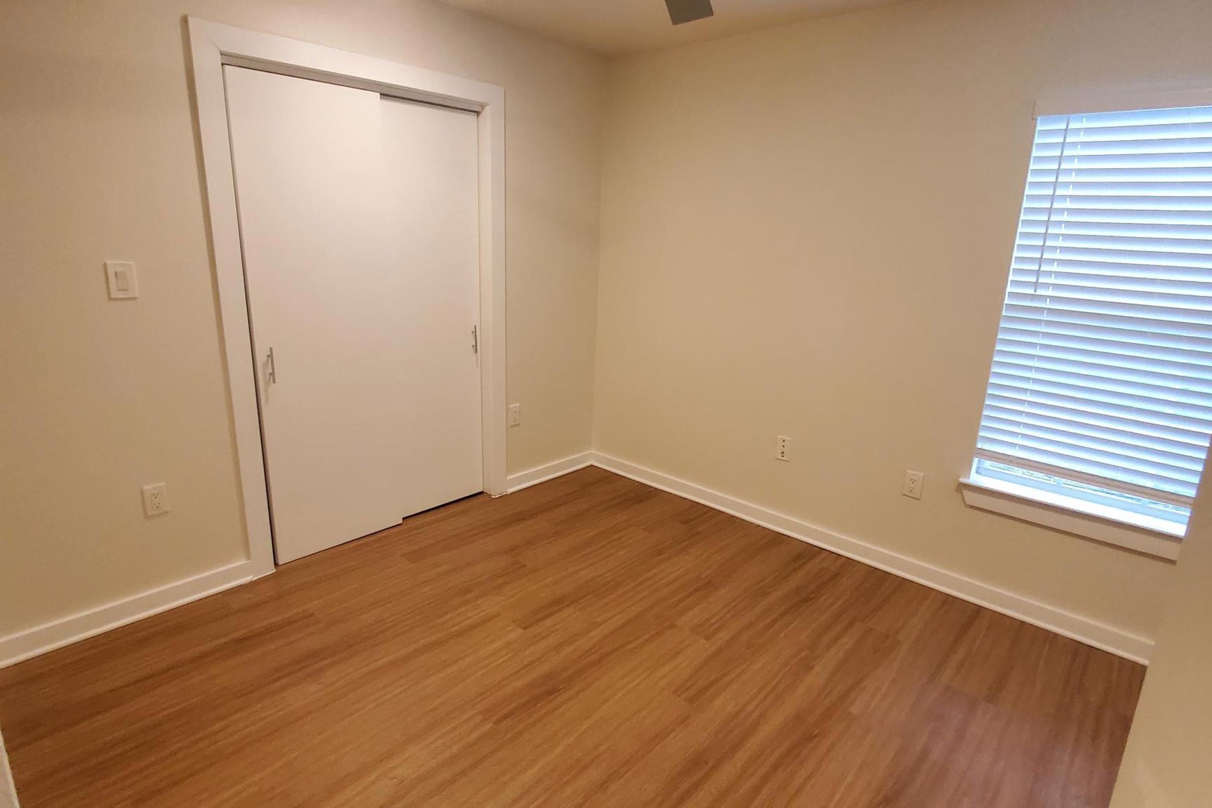 A small, empty room featuring light-colored walls, a wood laminate floor, and a window with blinds. There is a closet with sliding white doors on one wall, and the room is well-lit with a ceiling fan visible.