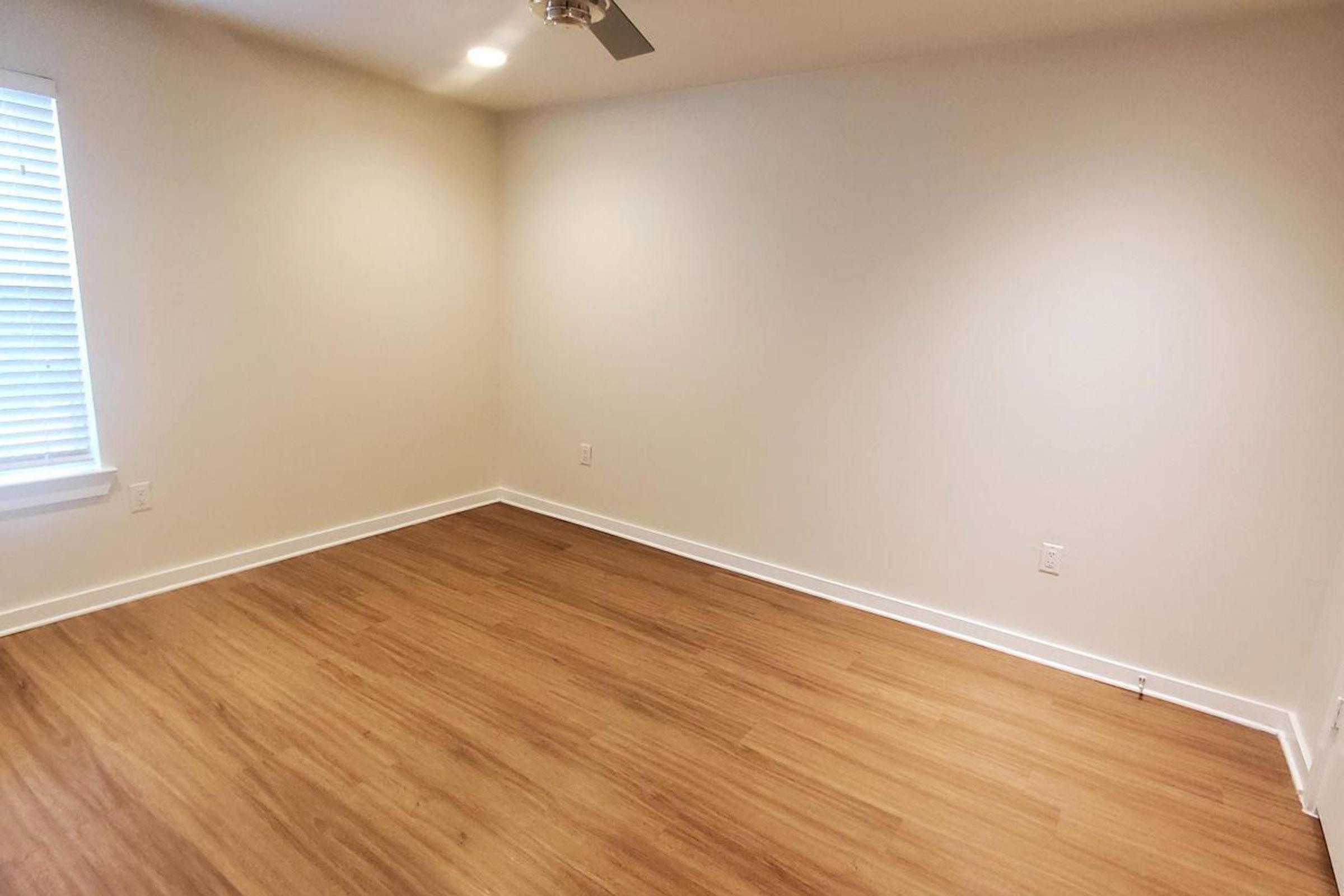 A vacant room with light-colored walls and a wooden floor. A ceiling fan is visible, and there is a window with white blinds allowing natural light to enter. The space is minimalistic, emphasizing its emptiness and potential for decoration or furnishing.