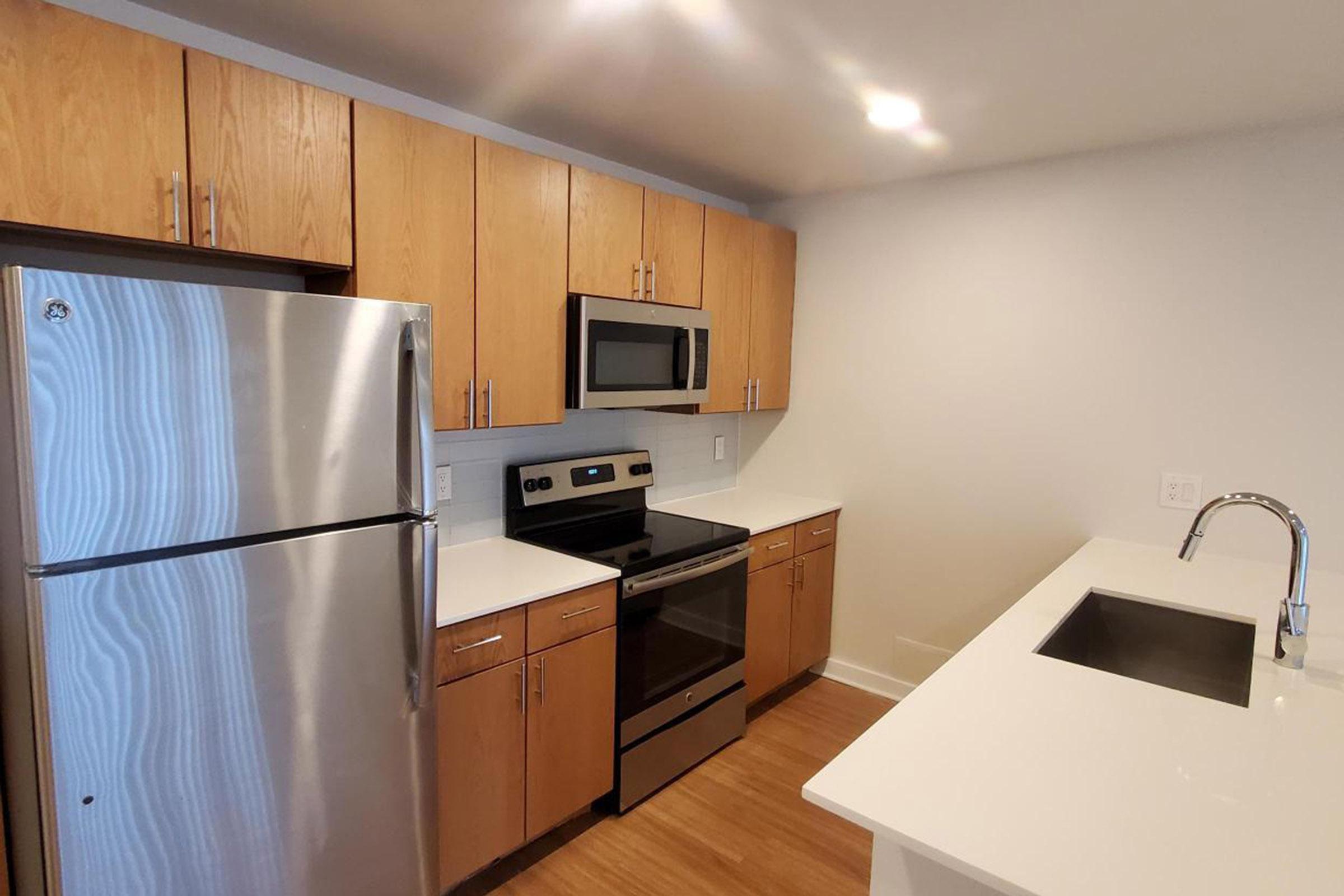 Modern kitchen featuring stainless steel appliances, including a refrigerator, microwave, and oven. The cabinetry is made of light wood, and the countertops are white. There's a sink on an island with a sleek faucet, and the flooring is a light hardwood. Bright lighting highlights the contemporary design.