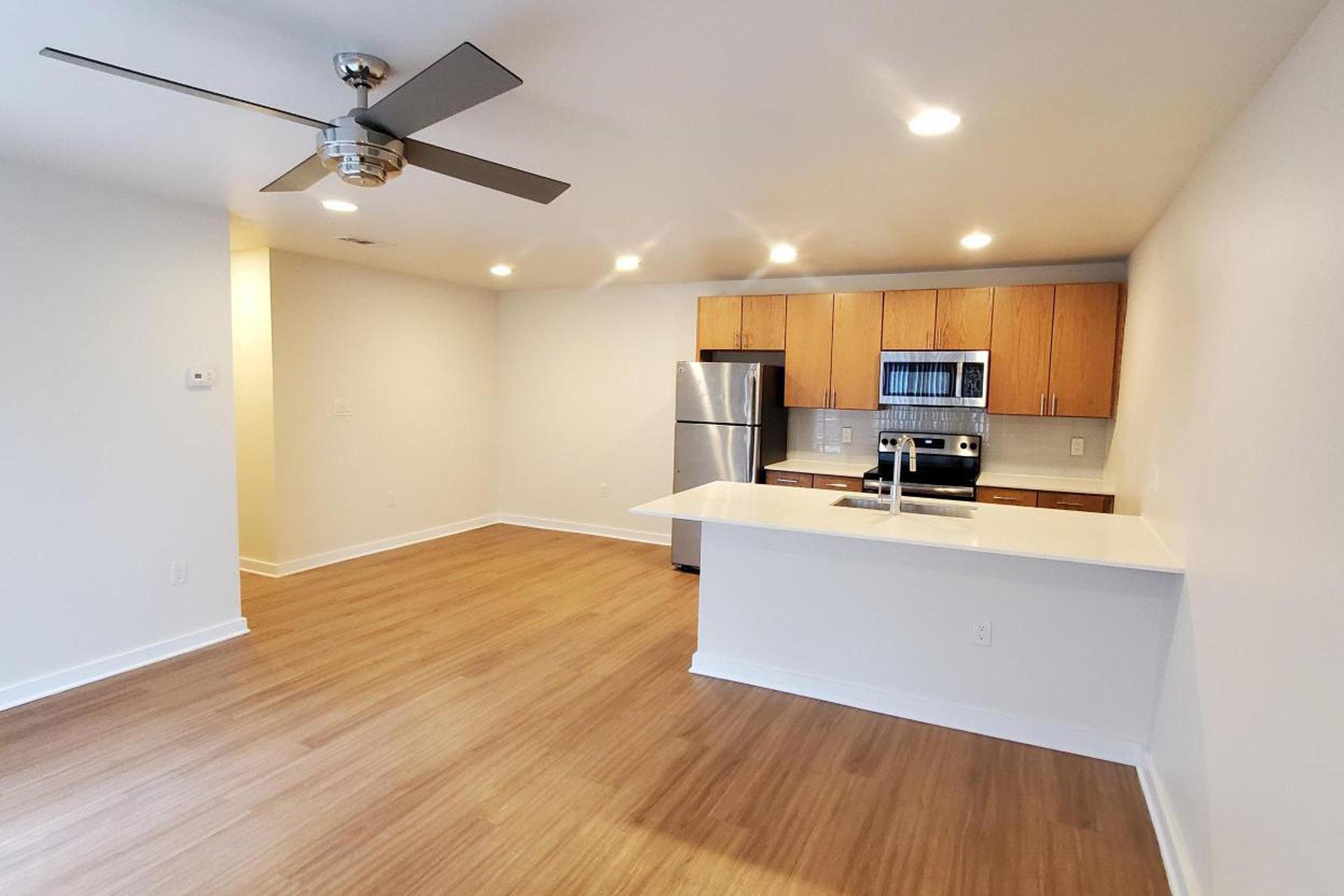 Modern kitchen and living area with light wood flooring, featuring a ceiling fan, stainless steel appliances, and wooden cabinets. The space is well-lit with natural and artificial light, showcasing an open layout that connects the kitchen to the living room.
