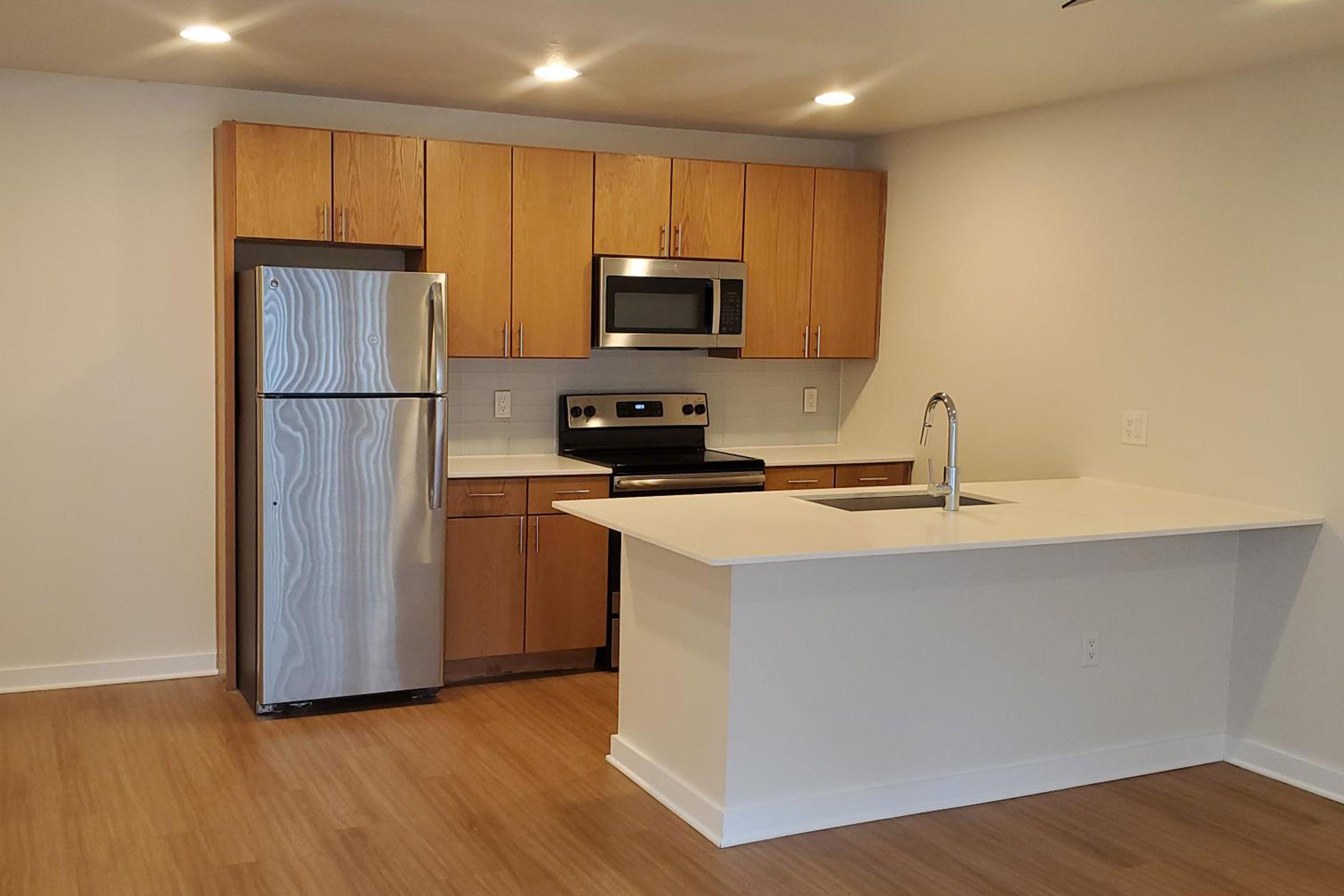 Modern kitchen featuring stainless steel appliances, including a refrigerator and microwave. The cabinets are light wood, and the countertops are white. A central island with a sink is present, and the flooring is a warm wood finish. The space is well-lit with recessed lighting.