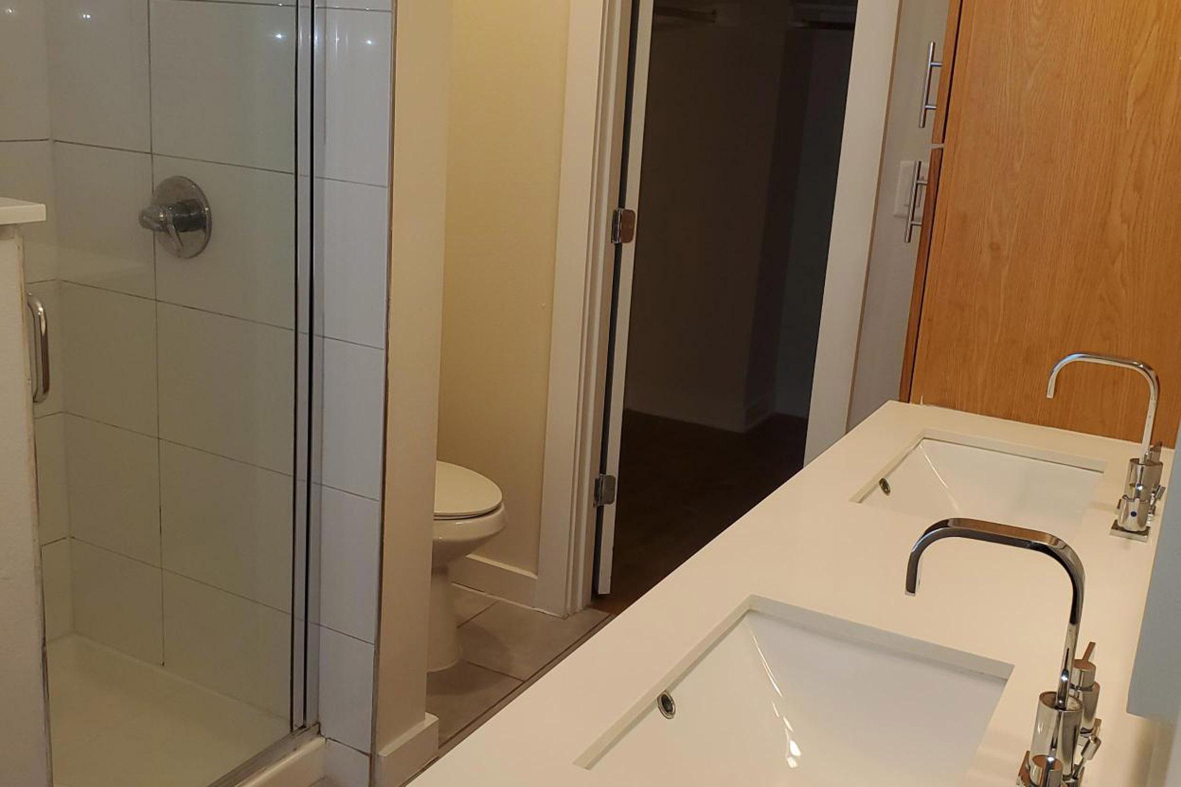 A modern bathroom featuring a glass shower enclosure, a white tiled floor, and a double sink vanity with sleek faucet fixtures. A toilet is visible in the background, along with a door leading to a closet or storage area. The overall design is clean and contemporary.