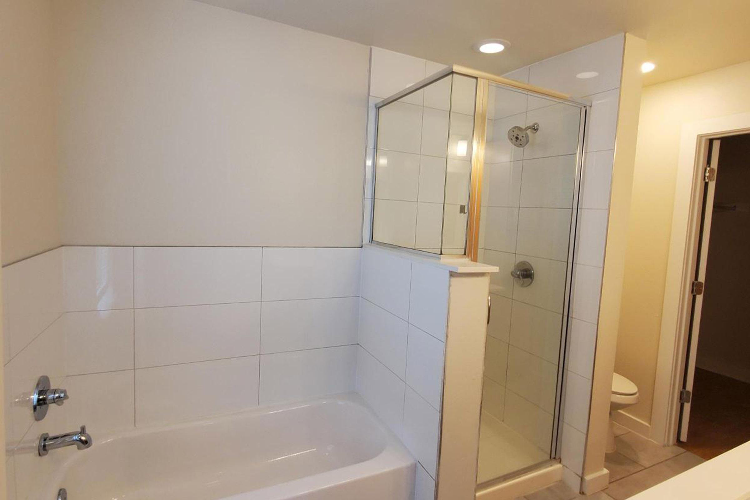 A modern bathroom featuring a glass shower enclosure, a white bathtub, and tiled walls. The lighting is bright, and there are clean lines and minimal decor, creating a spacious and inviting feel. A toilet is partially visible in an adjacent room.