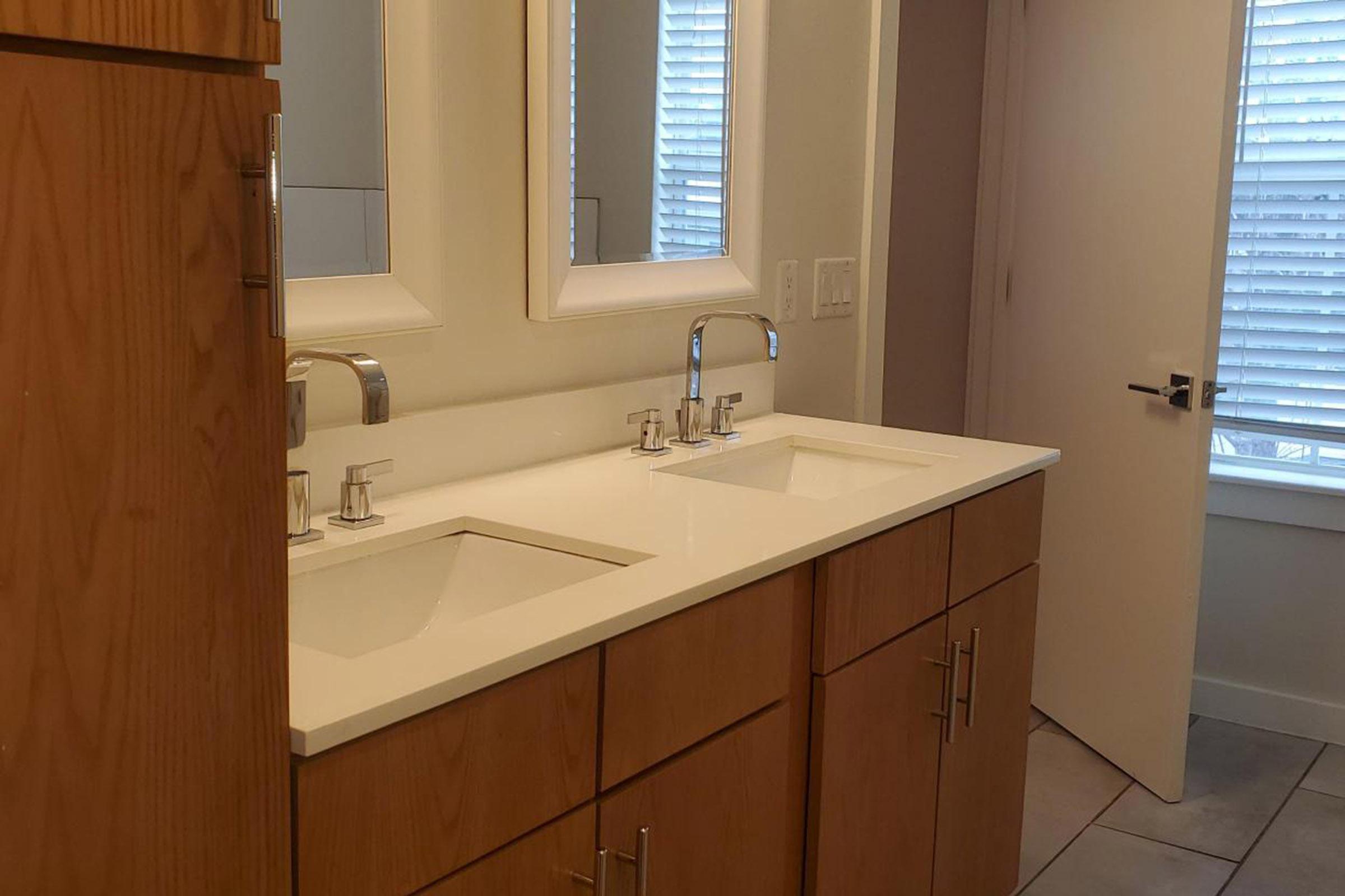 A modern bathroom with a double vanity featuring two sinks and stylish faucets. The cabinetry is light wood, and there are two rectangular mirrors above the sinks. A door is visible on the right, leading to another room, with natural light entering through a window with blinds.