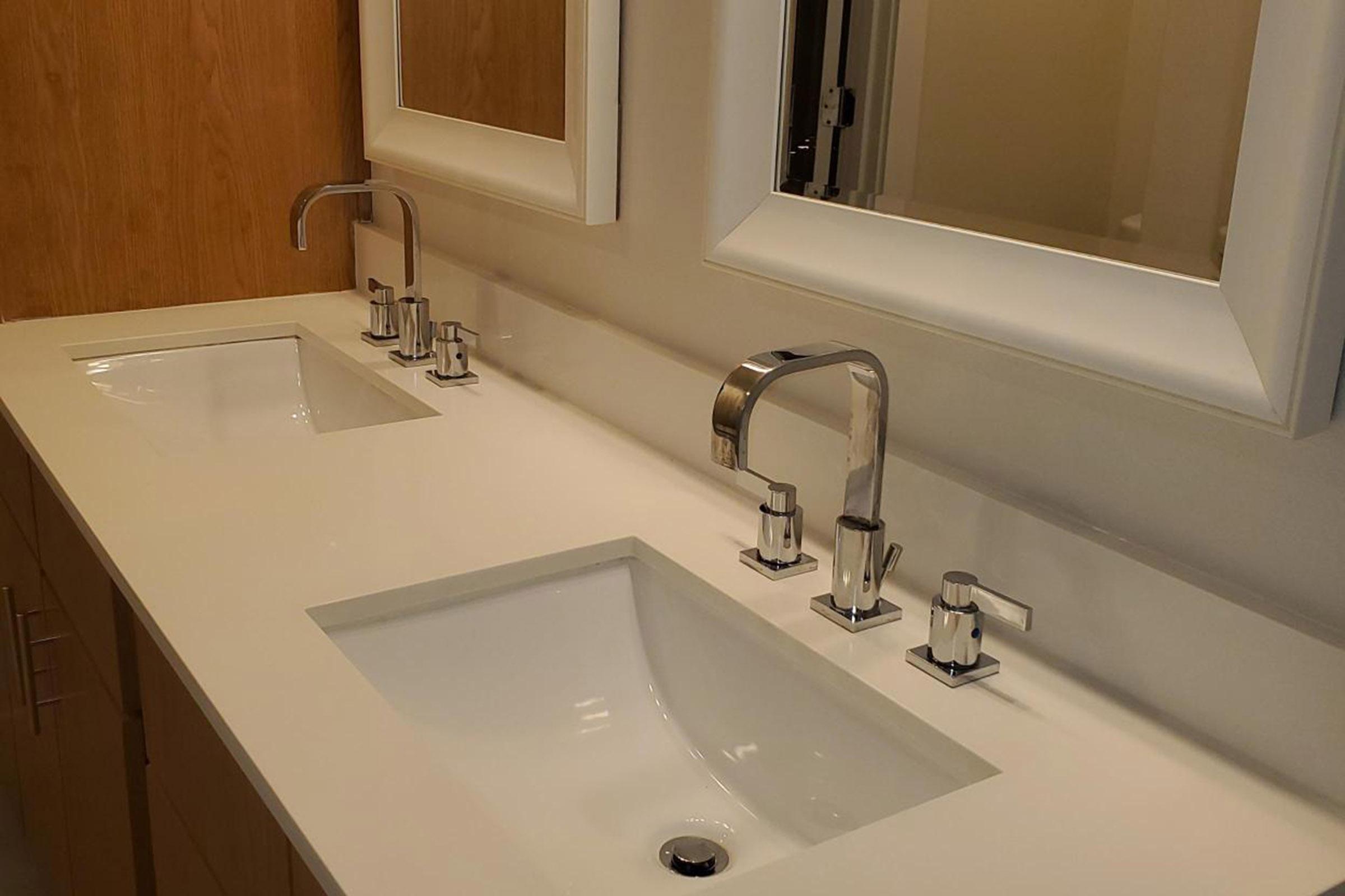 A modern bathroom with a minimalist design featuring a double sink countertop. Each sink has a sleek, silver faucet, and there are two rectangular mirrors above the sinks. The walls are light-colored, and the cabinetry is made of a warm wood finish, creating a clean and stylish atmosphere.