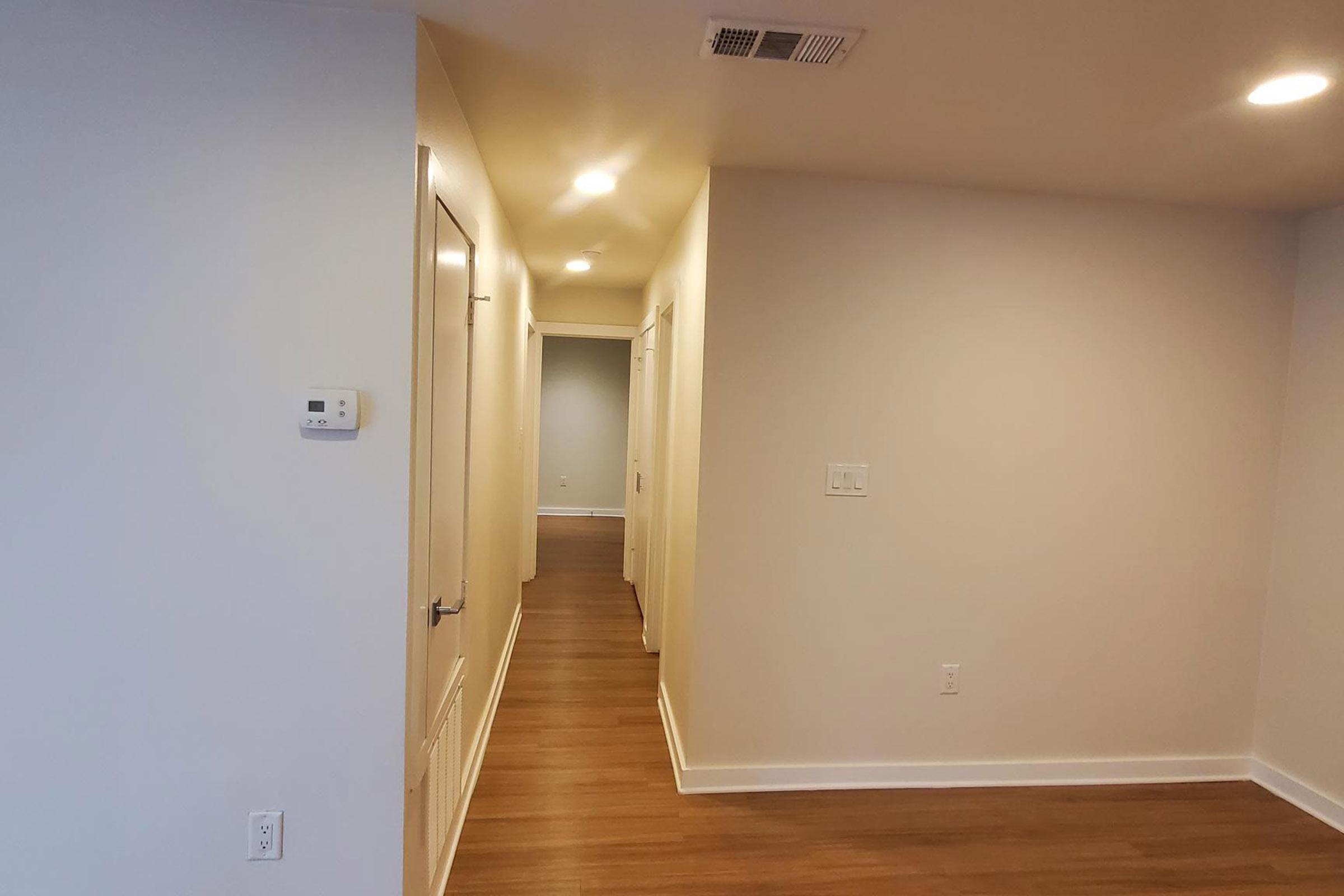 A well-lit hallway featuring smooth walls and a light wooden floor. There are doors on either side, and the area is illuminated by ceiling lights. The space has a clean, modern appearance with a neutral color palette.