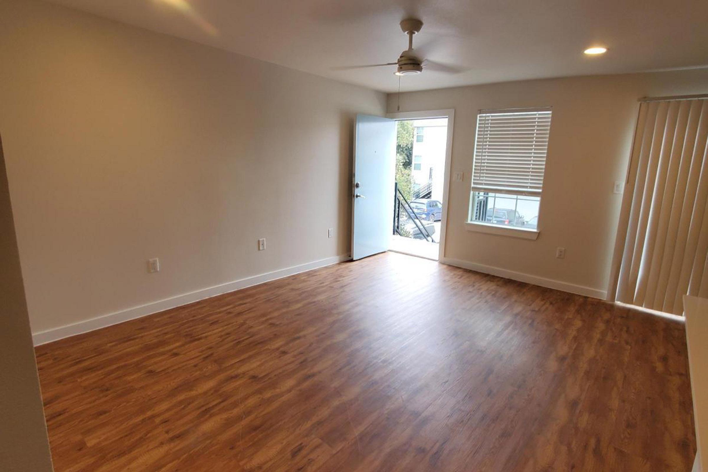 A spacious, empty living room with light-colored walls and wooden flooring. A ceiling fan is visible, and a door leads outside, with a window featuring blinds. The room is well-lit and has a modern feel, ideal for furnishing and personalizing.