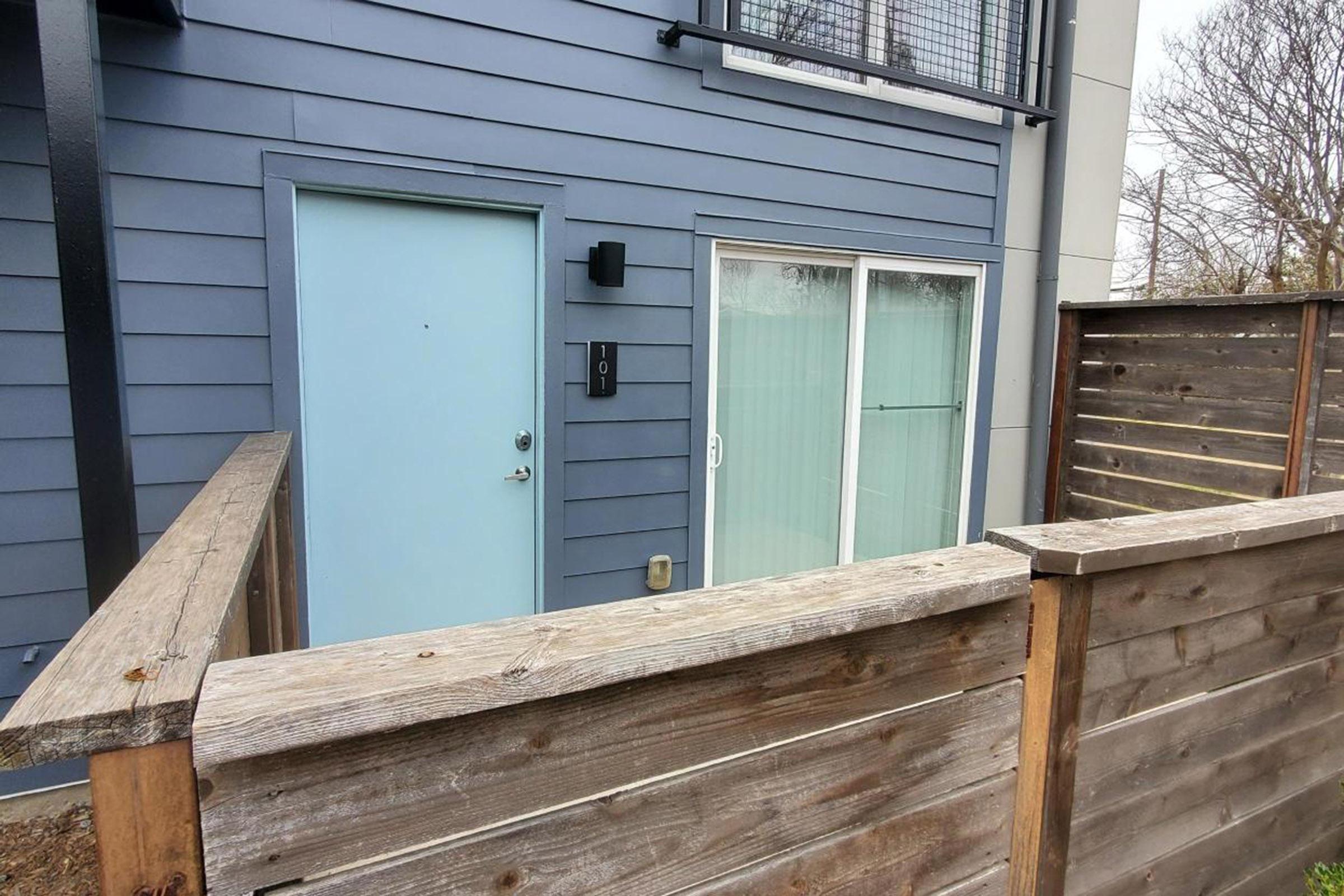 A blue exterior wall of a house featuring a light blue door, a wall-mounted intercom system, and a large window with sheer curtains. A wooden fence encloses the area, providing privacy and separation from the surrounding environment.