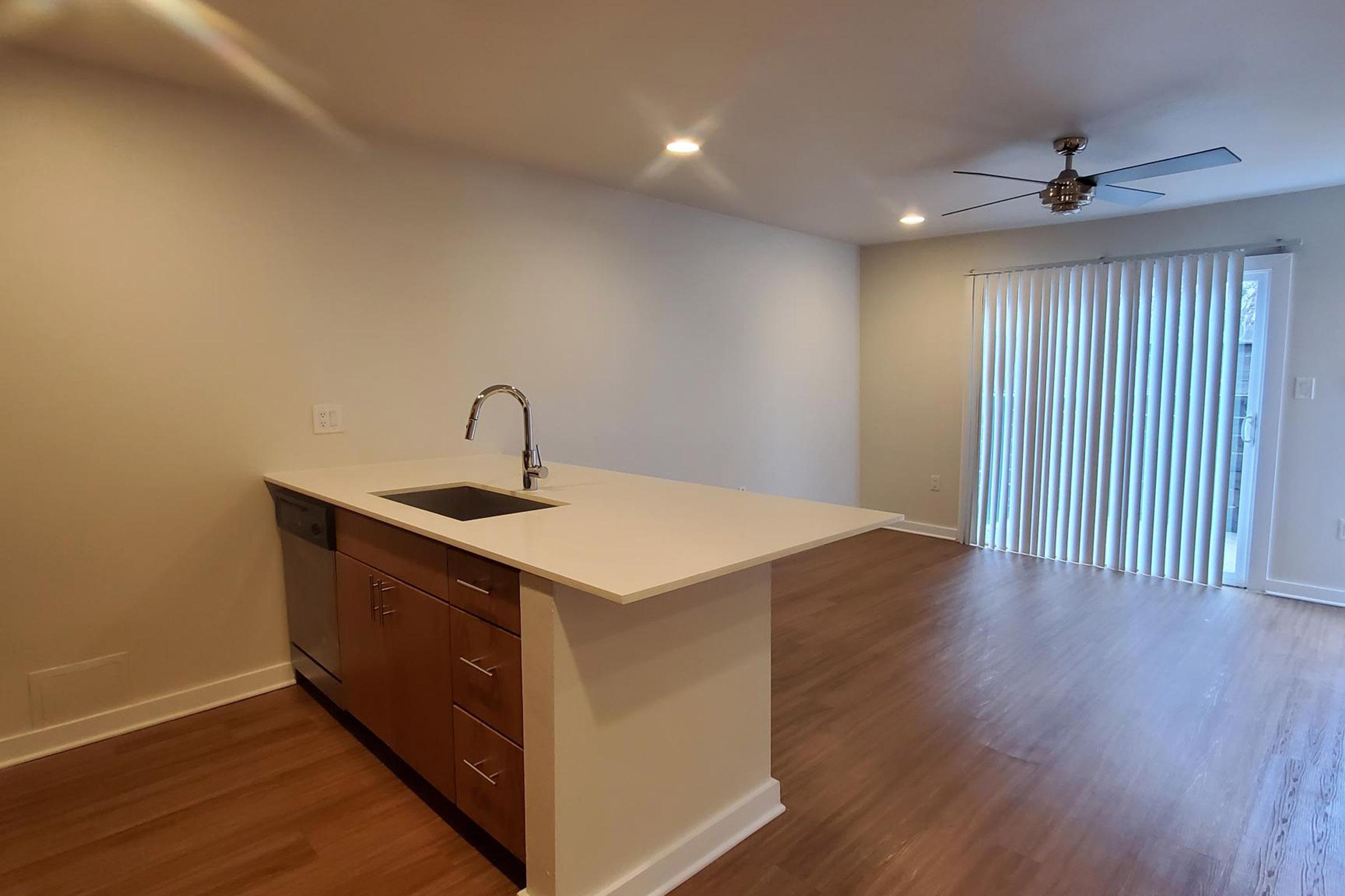 Spacious modern kitchen with a white countertop and stainless steel sink, featuring wooden cabinets. Bright open living area with sliding glass door leading to an outdoor patio, natural light streaming in from large windows, and a ceiling fan for ventilation. Hardwood floors throughout.
