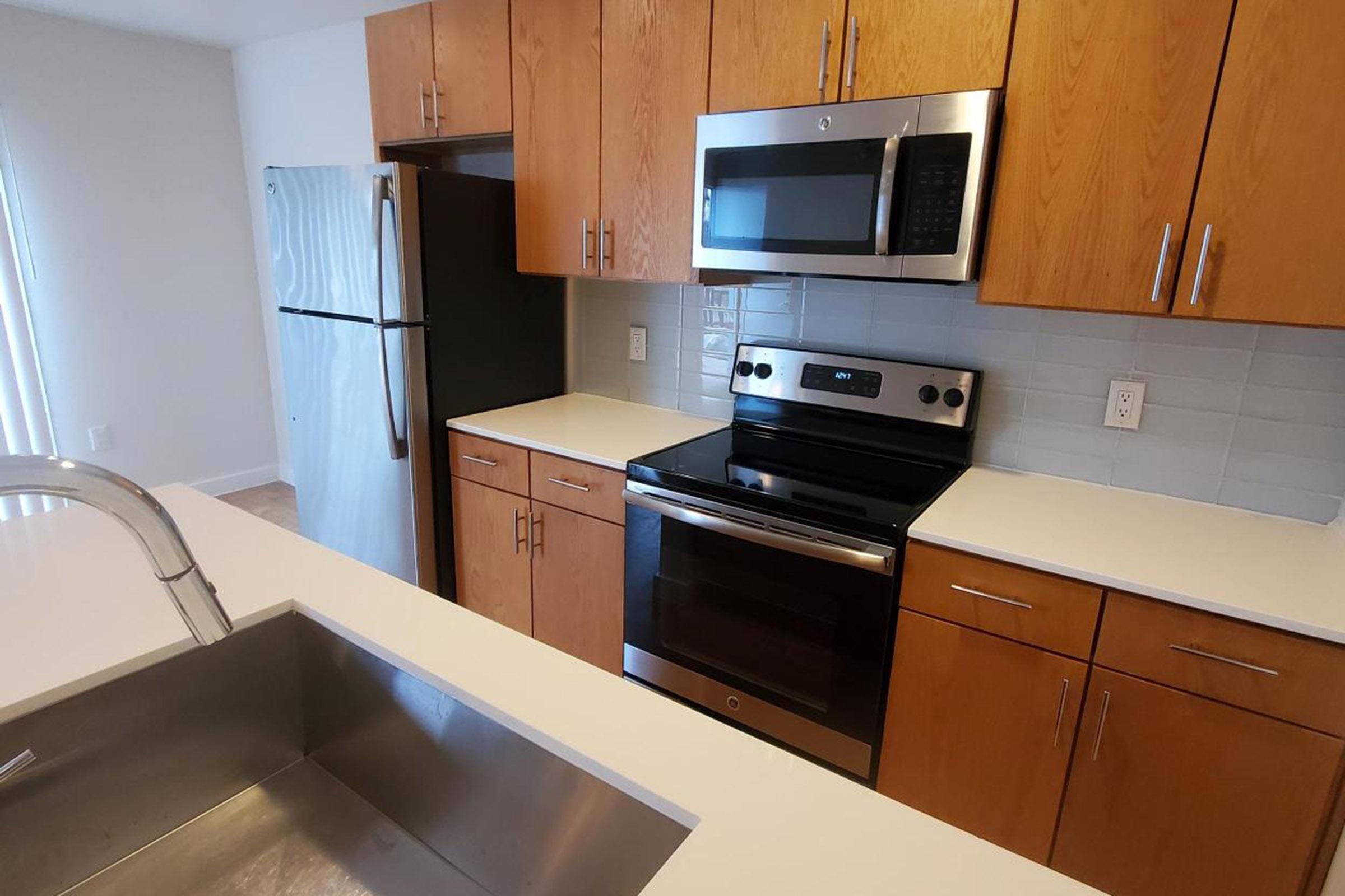 Modern kitchen featuring wooden cabinetry, stainless steel appliances including a refrigerator, microwave, and oven, with a white countertop and a large sink. The kitchen has bright, natural lighting and a minimalist design.