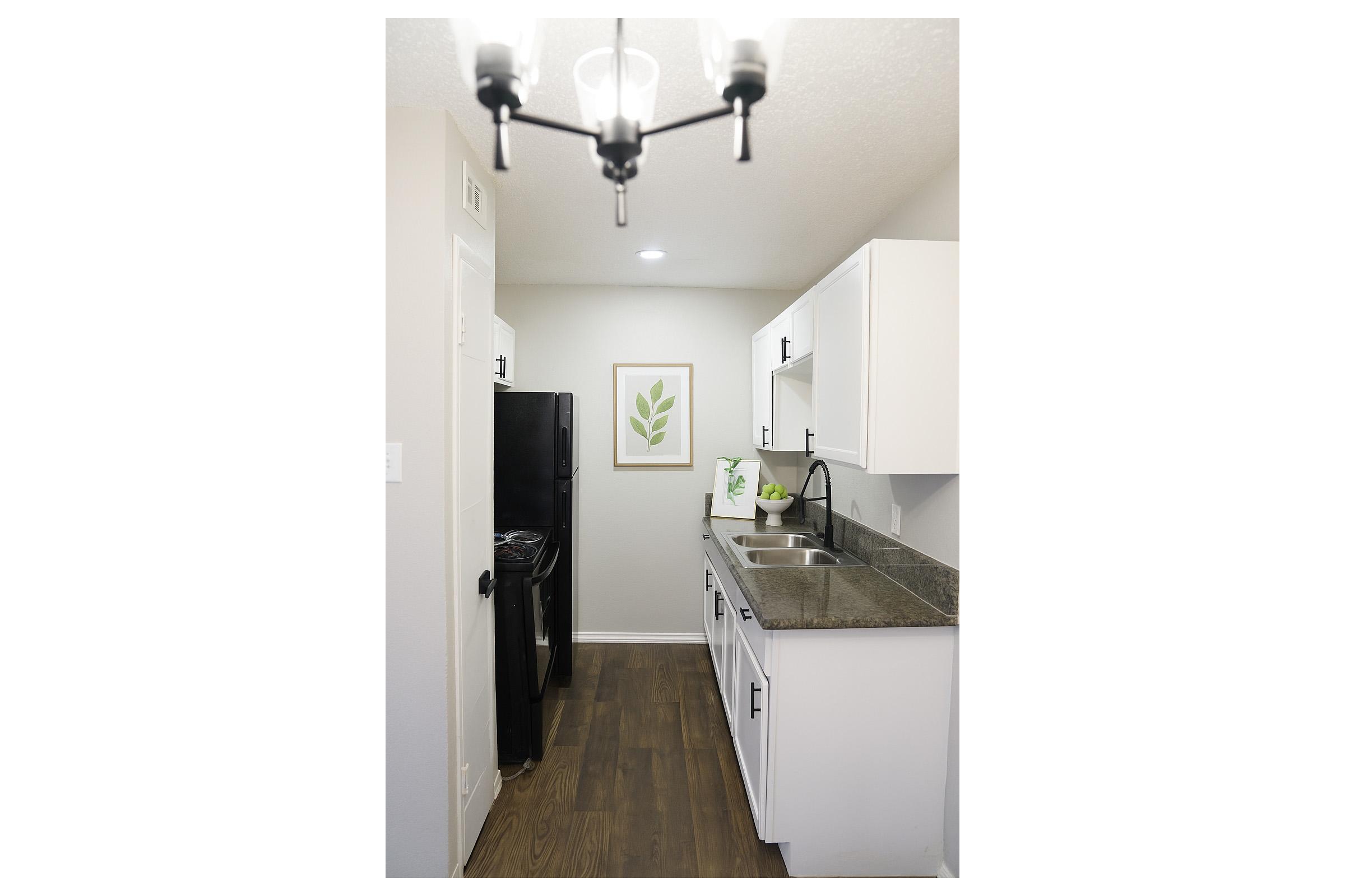 A modern kitchen featuring white cabinets and dark countertops. There is a black refrigerator and a sink with a faucet. The walls are light-colored, and a framed green leaf artwork is displayed on one wall. Natural light illuminates the space, creating a bright and inviting atmosphere.