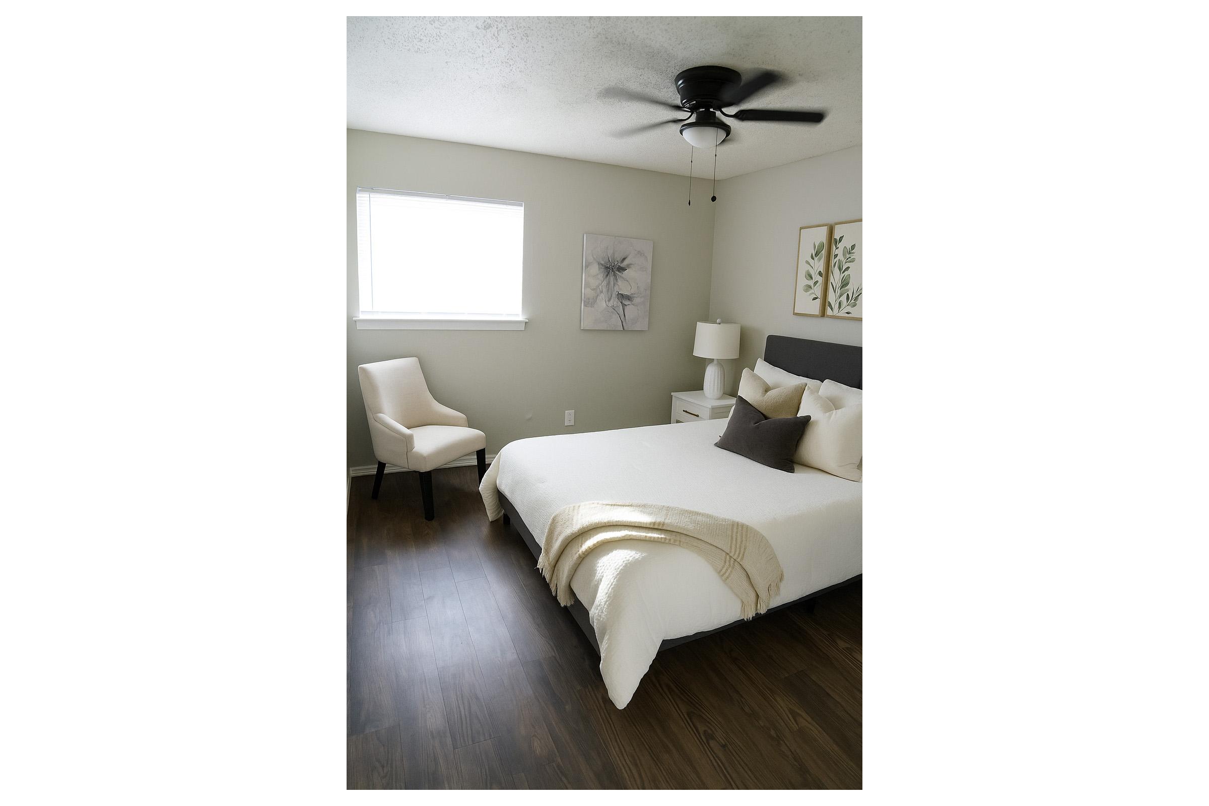 A cozy bedroom featuring a white bed with neutral bedding, a dark headboard, and two decorative pillows. There is a white chair next to the bed, a lamp on a nightstand, and artwork displayed on the wall beside a window. The floor is dark wood, creating a warm atmosphere.