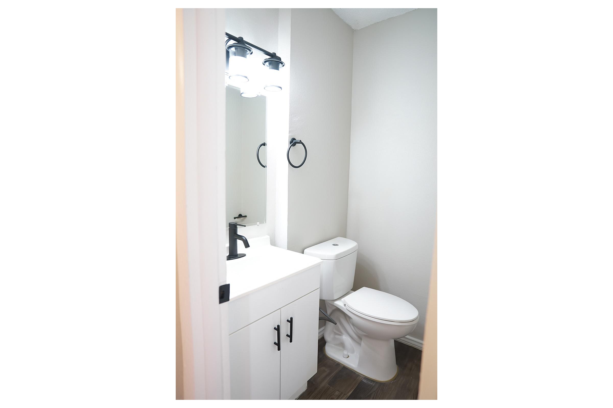 A small modern bathroom featuring a white vanity with a sink, a mirror with lights above, a round mirror on the wall, a toilet, and neutral-colored walls. The flooring is a dark wood-like material, creating a contemporary look.