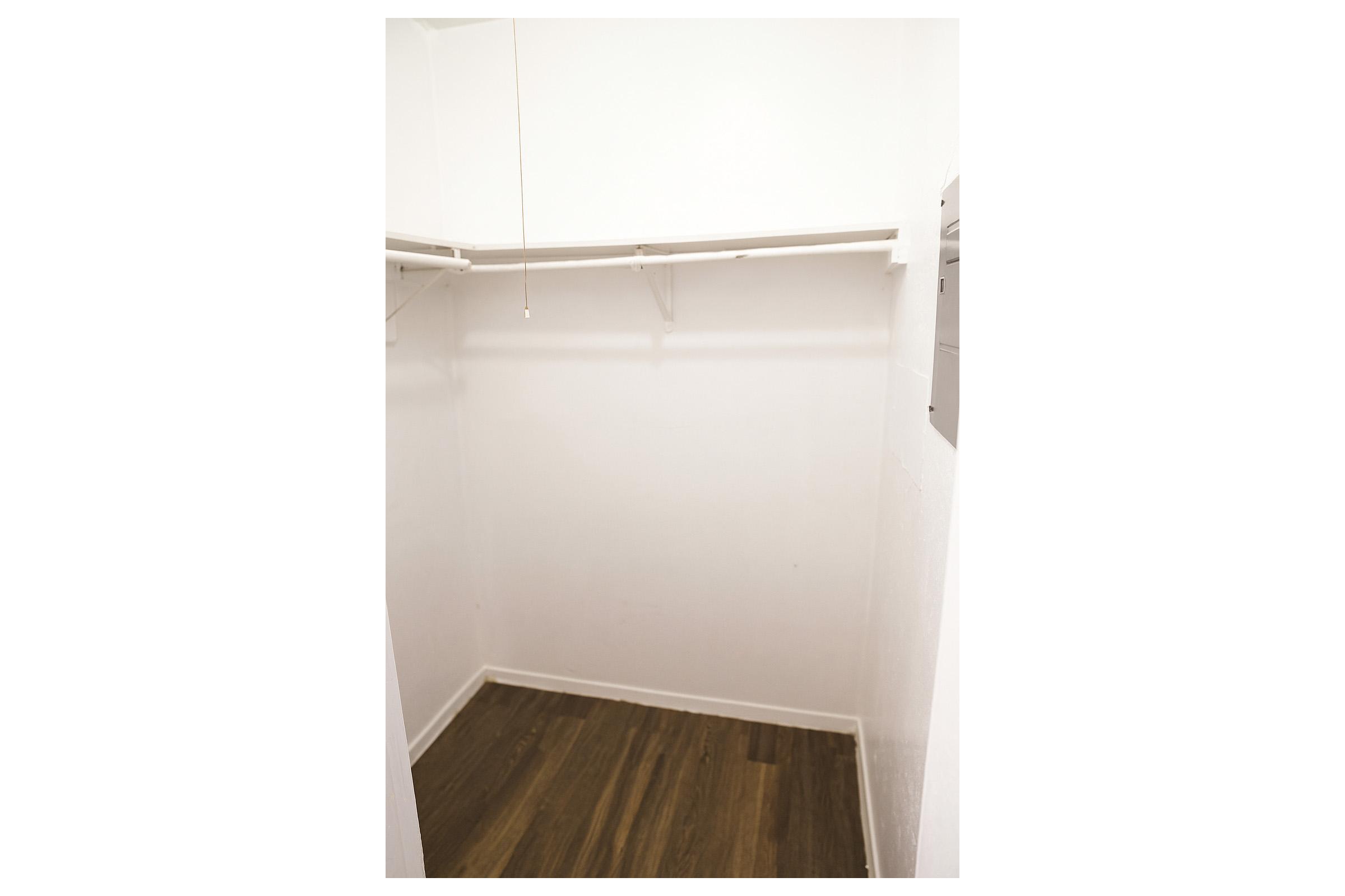 Empty closet with white walls and a wooden floor. There are two shelves along the upper wall and a light fixture hanging from the ceiling. The space is uncluttered and minimalistic, providing a clear view of the interior.