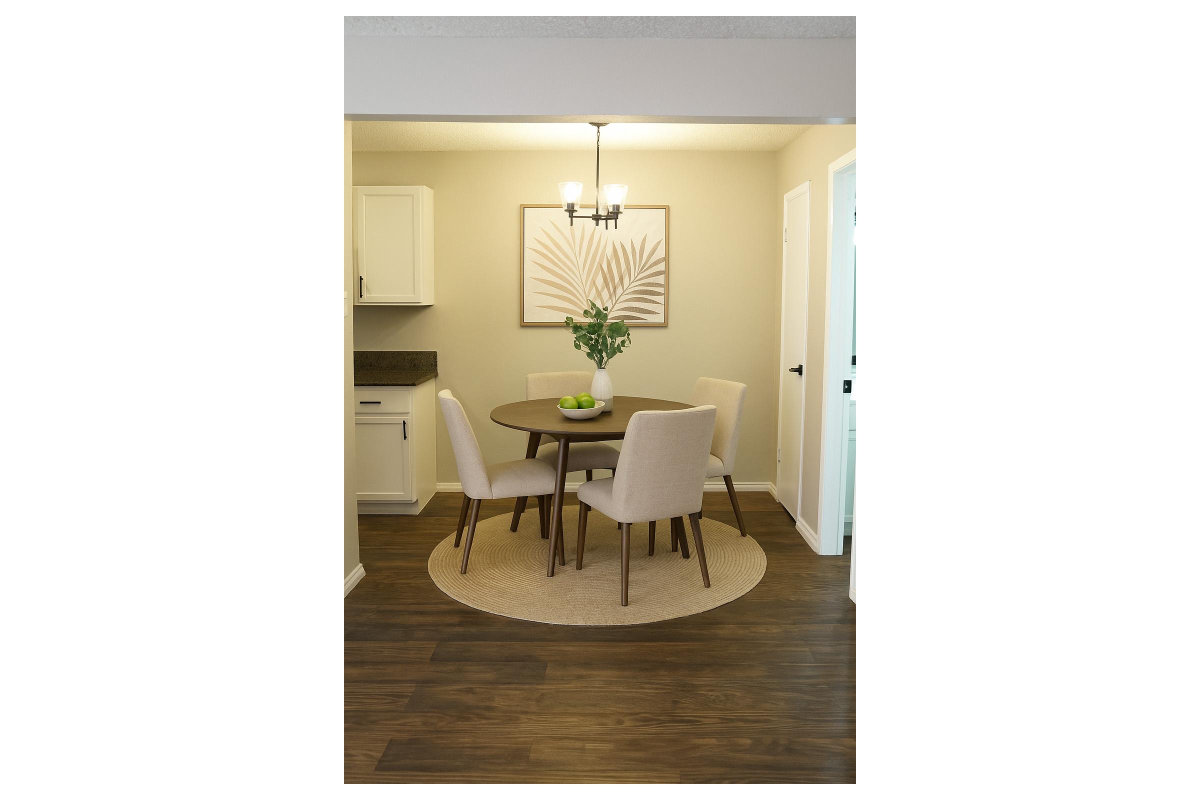 A cozy dining area featuring a round wooden table with four light-colored upholstered chairs. A centerpiece of green apples sits on the table. The room is illuminated by a stylish chandelier above, and a decorative wall art piece with a leaf design enhances the ambiance. Natural wood flooring and neutral walls complete the scene.