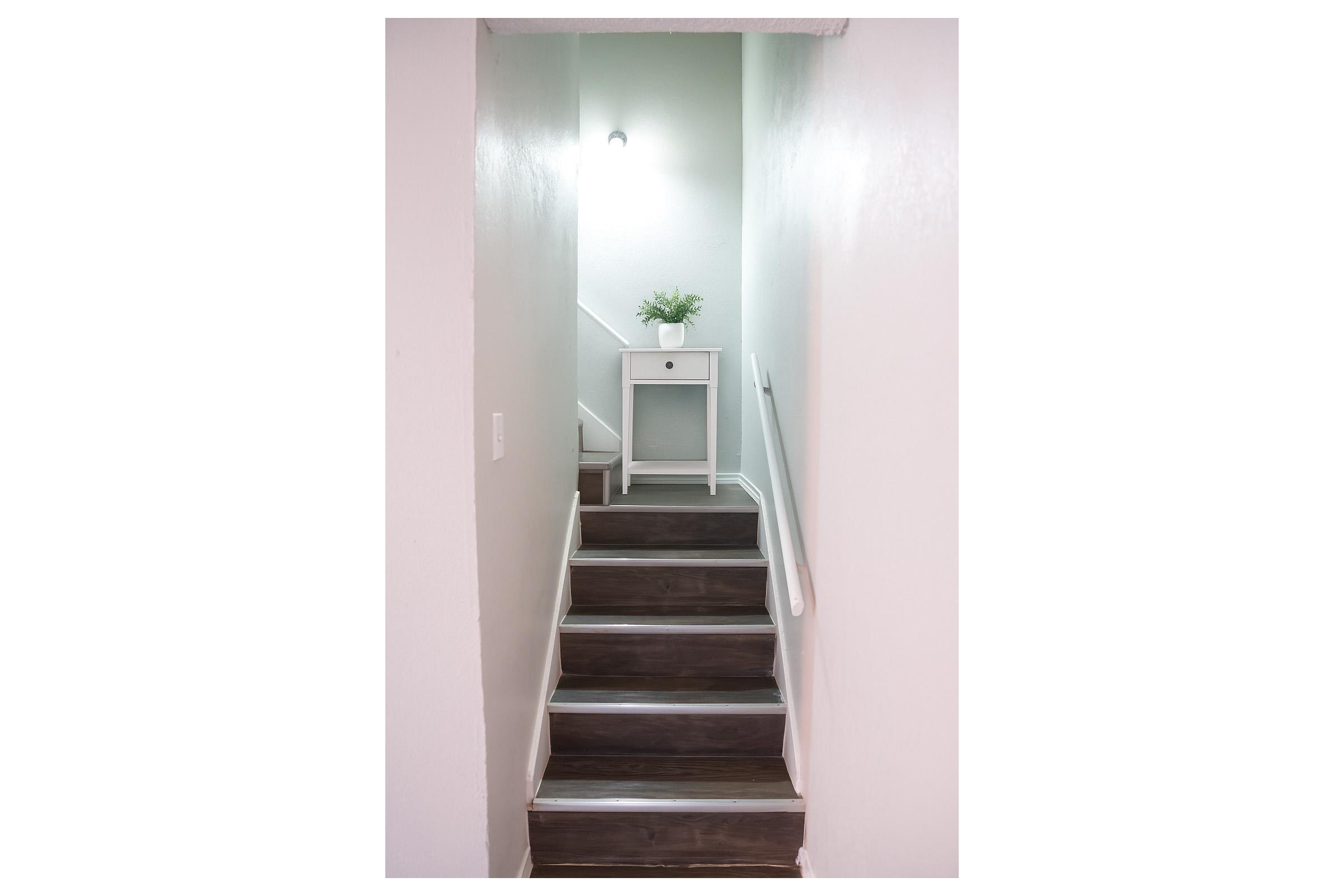 A view of a stairway leading to an upper level, featuring wooden steps and a light-colored wall. At the top of the stairs, there is a small white side table with a potted plant, illuminated by a wall-mounted light. The overall atmosphere is bright and inviting.