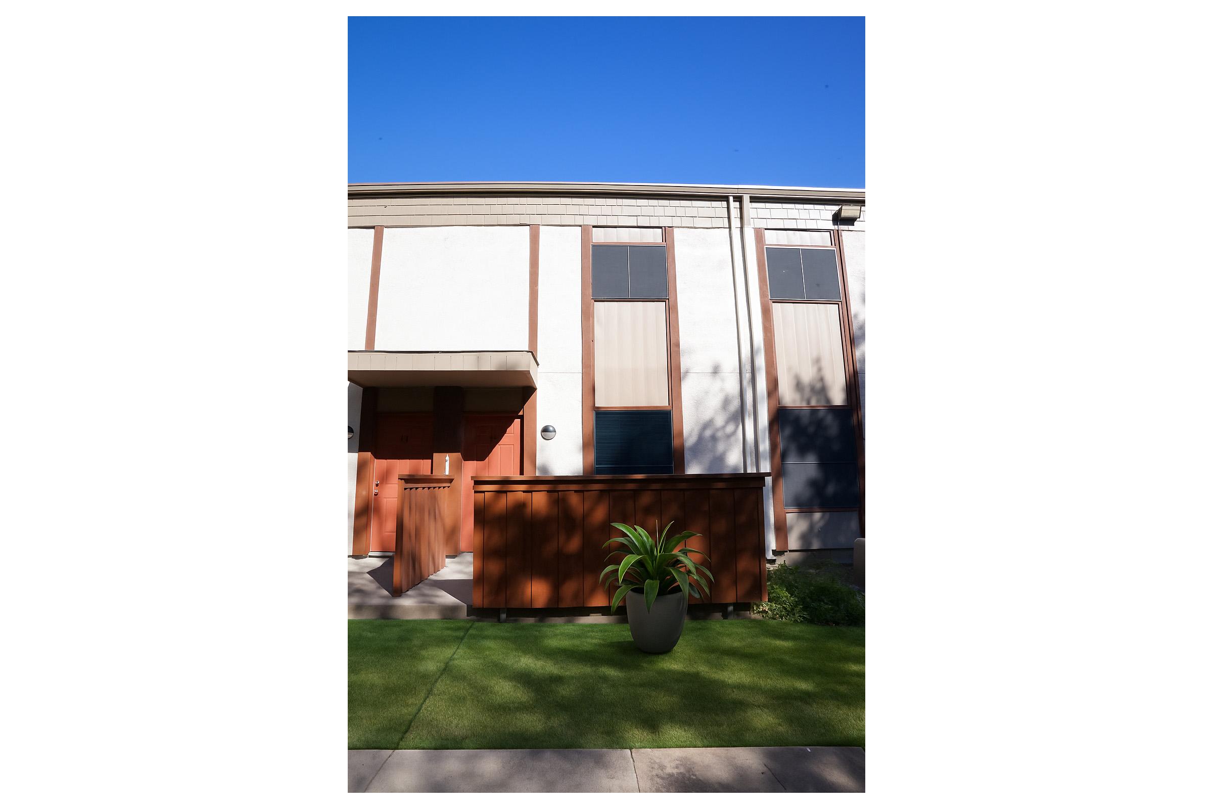 A modern building facade featuring a wooden door with an adjacent planter. The structure has large windows and a minimalist design, set against a clear blue sky. Lush greenery and artificial grass complete the outdoor area.
