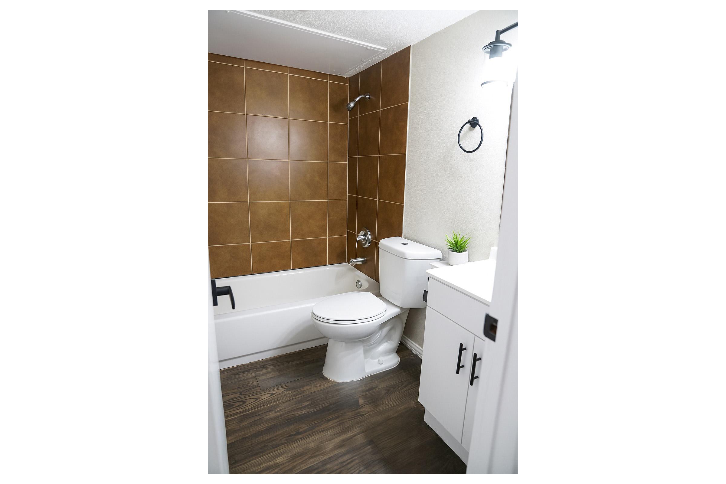 A clean, modern bathroom featuring a white toilet, a beige bathtub with a showerhead, and a brown tiled wall. There is a small green plant on the white vanity, which has dark handles, and the floor is a dark wood finish. A wall-mounted light fixture provides illumination.