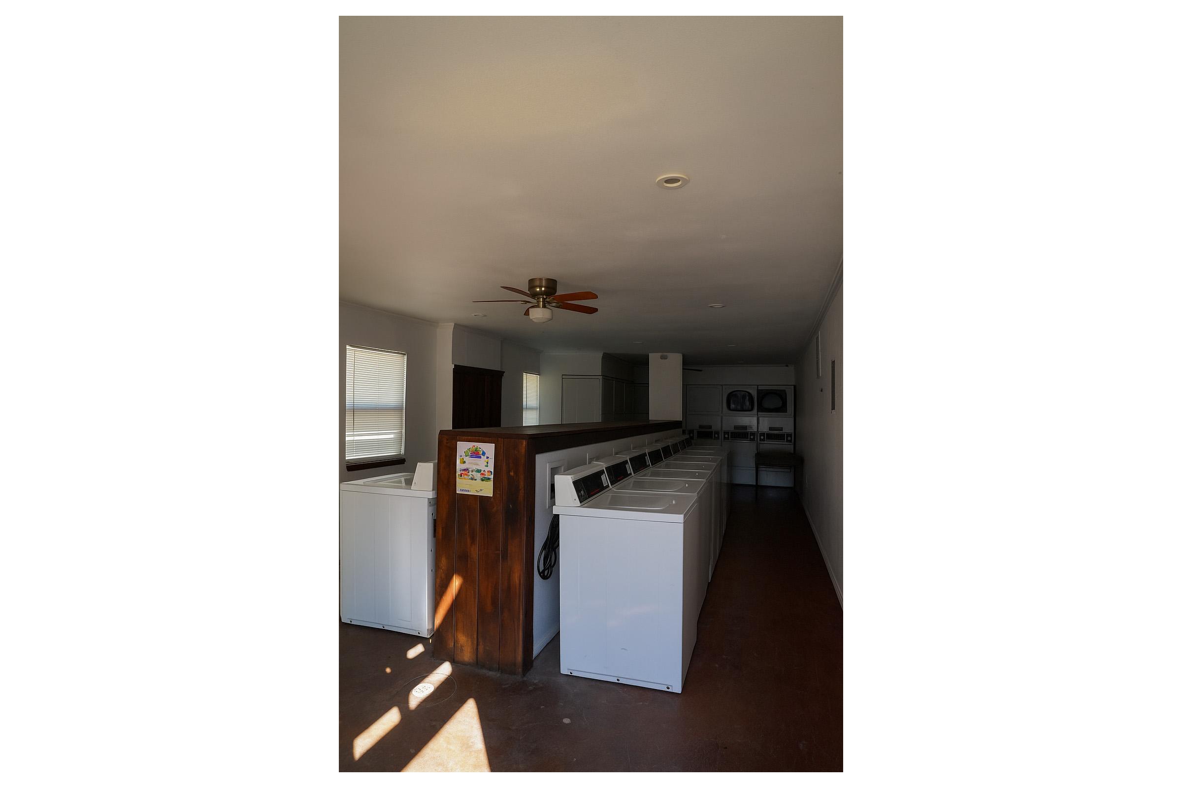 A clean, well-lit laundry room featuring several white washing machines lined up against a wooden partition. The room has a ceiling fan, windows allowing natural light, and additional laundry equipment visible in the background. The floor is brown and polished, contributing to the tidy appearance.