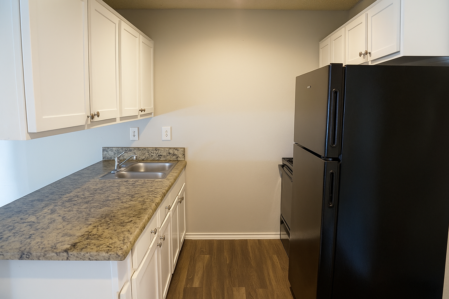 Interior view of a modern kitchen featuring white cabinets, a granite countertop, a stainless steel sink, and a black refrigerator. The walls are painted light gray, and the floor has a wood-like finish, creating a clean and contemporary look.