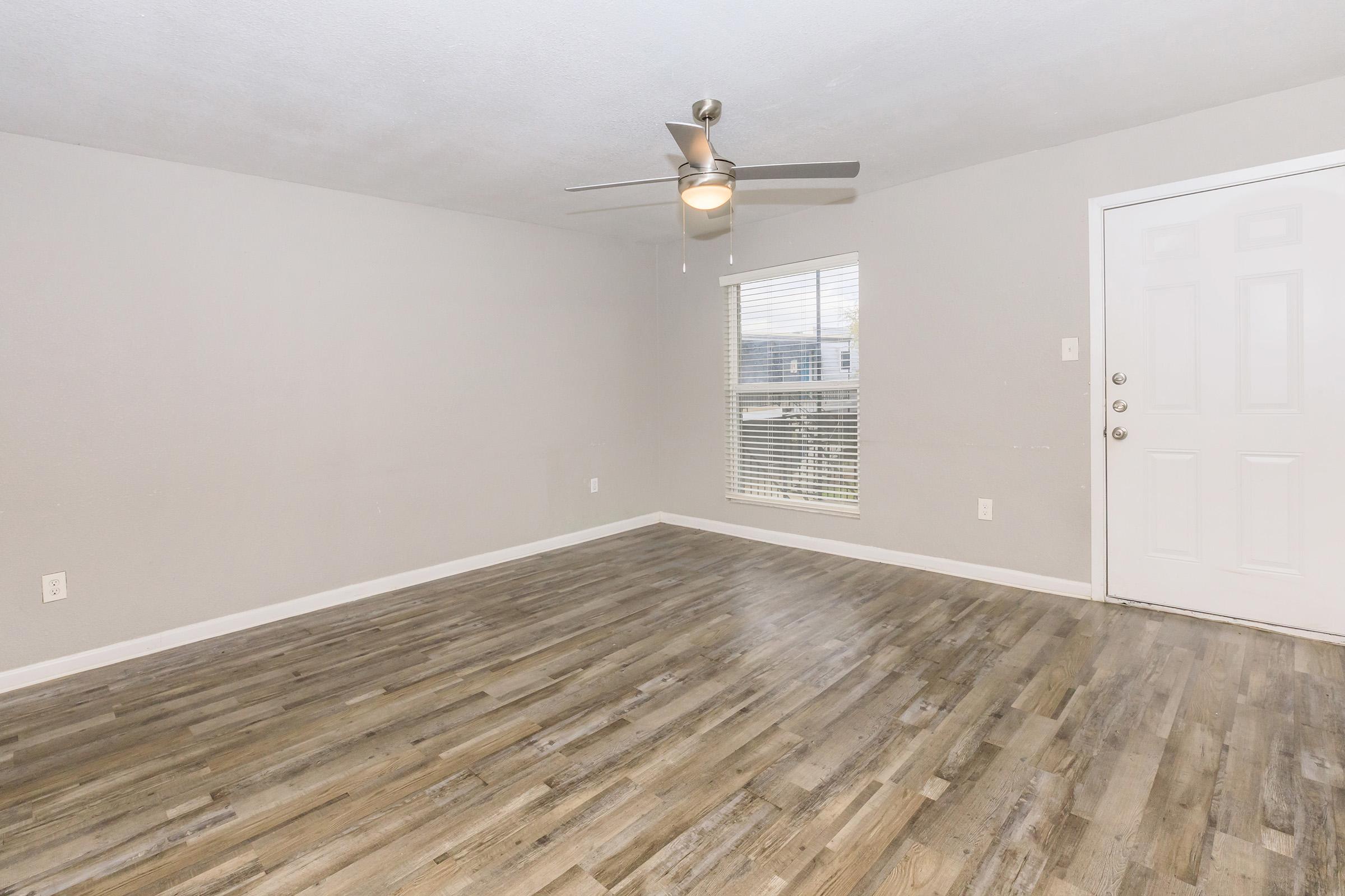 A spacious, empty room featuring light grey walls, a ceiling fan, and a large window with blinds. The floor has a wooden texture, and there is a white door visible on the right side of the image. The overall ambiance is bright and open, ideal for personalization or decoration.