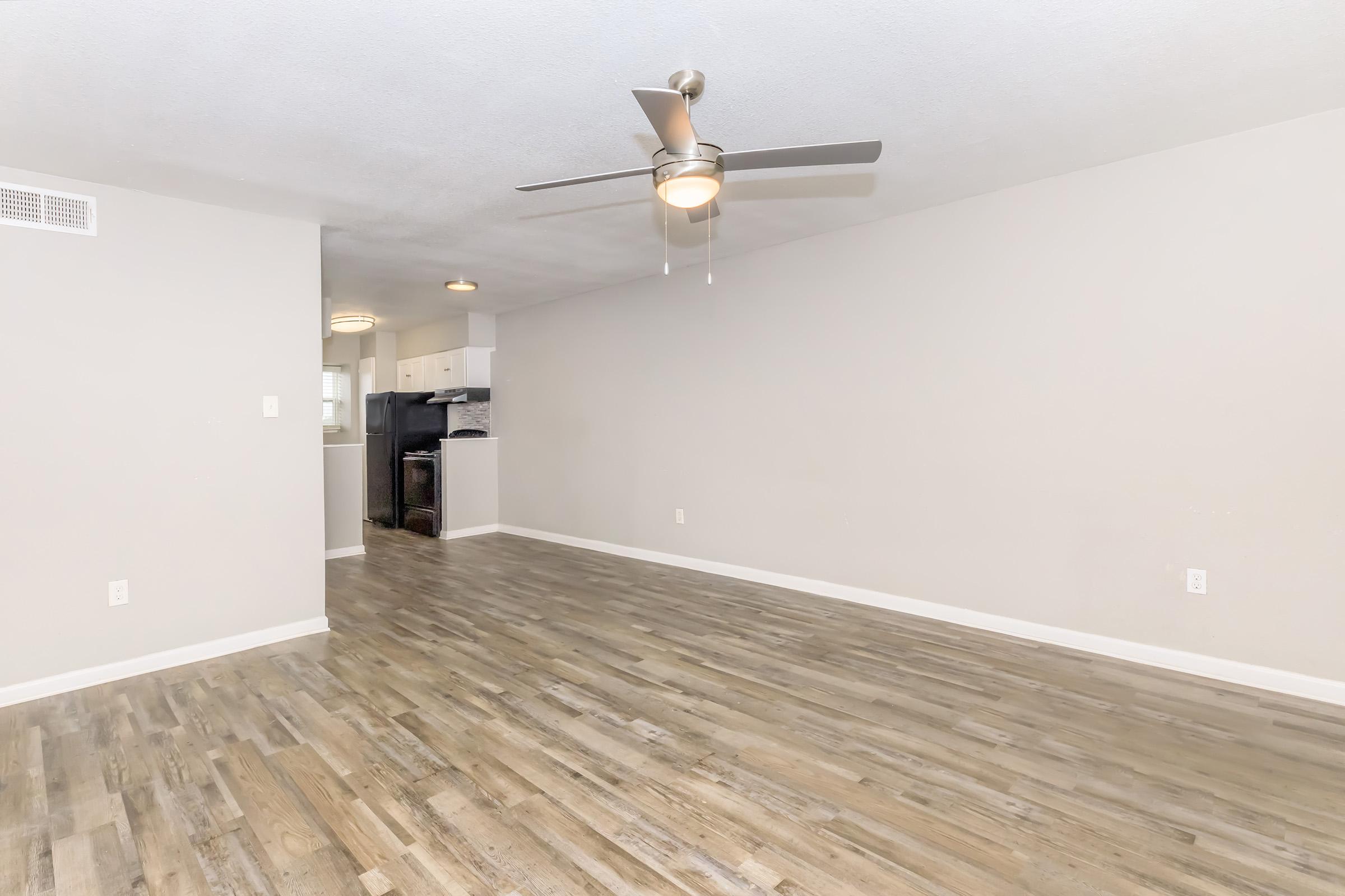 A spacious, modern living area featuring light-colored walls, a ceiling fan, and large wooden floors. The open layout leads into a kitchen area with white cabinets in the background, creating a bright and inviting atmosphere.