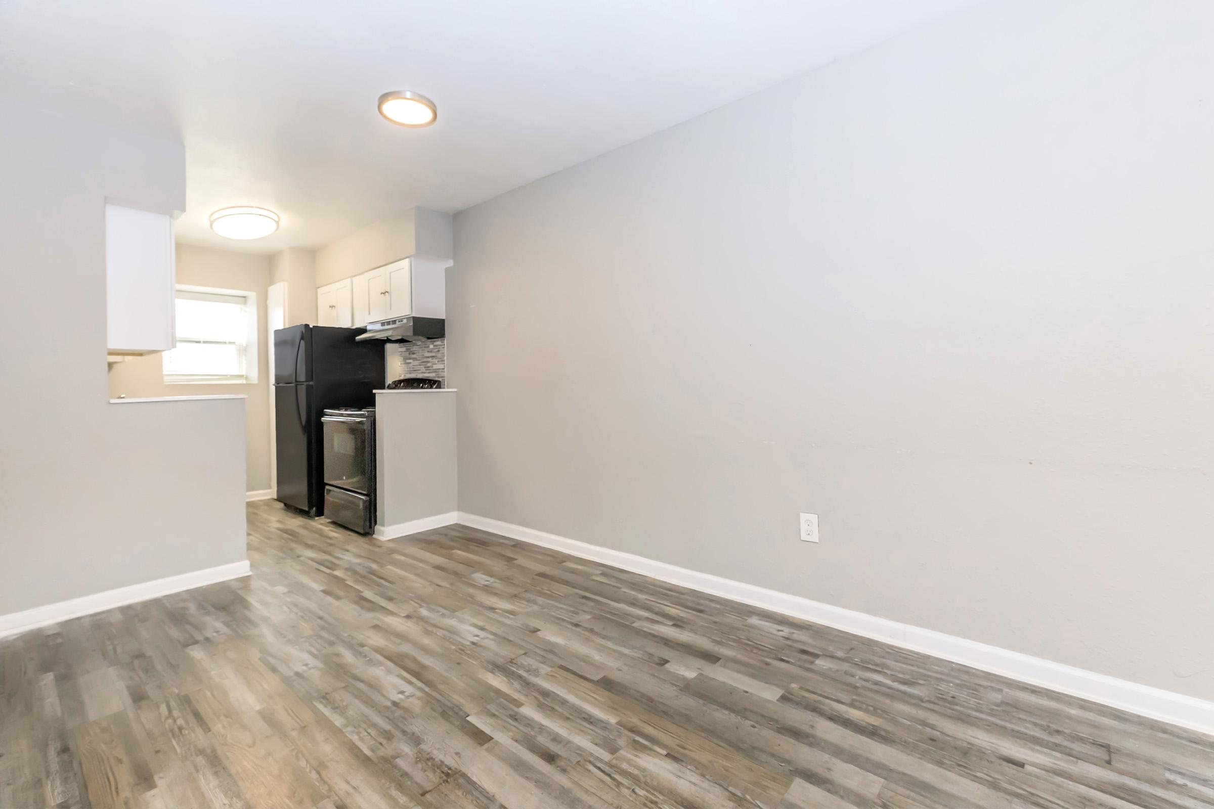 A spacious, well-lit kitchen area featuring a gray wall, light wood flooring, white cabinets, and a black refrigerator. The open layout connects to a living space, providing a modern and inviting atmosphere. Natural light from a circular ceiling light enhances the overall ambiance.