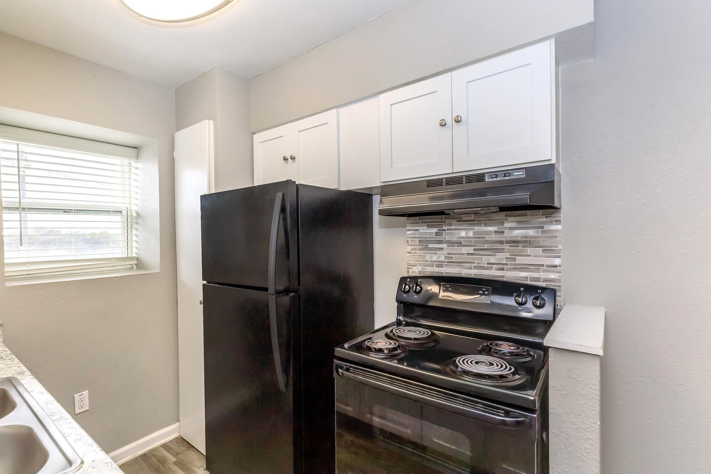 A modern kitchen featuring a black refrigerator and stove with a sleek design. White cabinets are located above, and the stove is accompanied by a silver hood vent. Light gray walls and a window allow natural light to brighten the space, complementing the overall contemporary aesthetic.