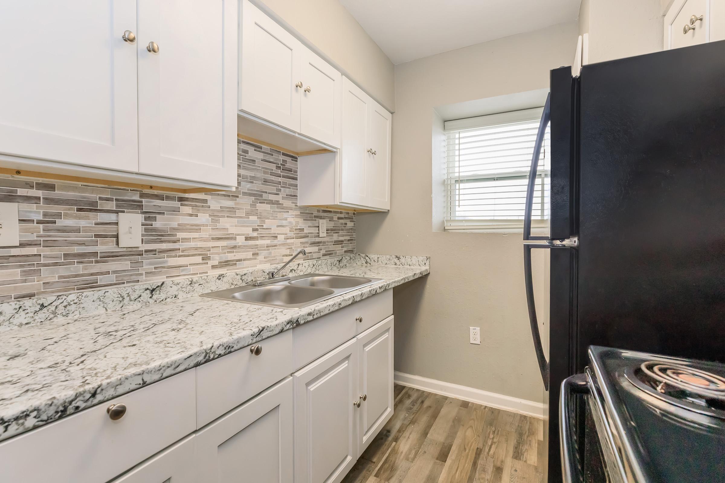 A modern kitchen featuring white cabinetry, a granite countertop, and a tiled backsplash. There is a stainless steel sink and black appliances, including a refrigerator and an oven. Natural light comes in through a window, enhancing the bright and clean ambiance of the space.