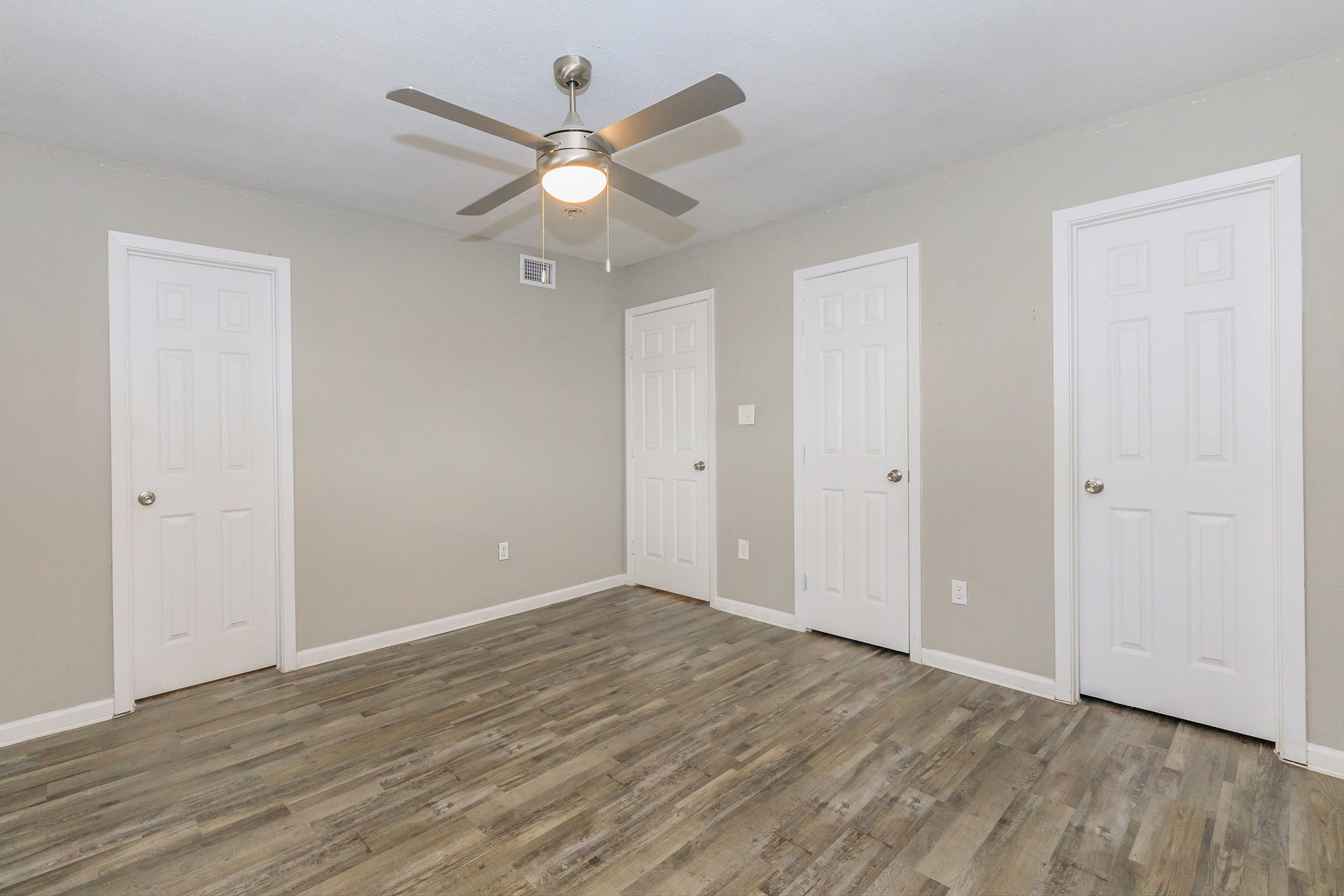 An empty room with light gray walls and a ceiling fan. The floor has wooden-like planks. Three closed white doors are visible on the walls, and there is a small light fixture in the center of the ceiling. The room has natural light and a clean, uncluttered appearance.