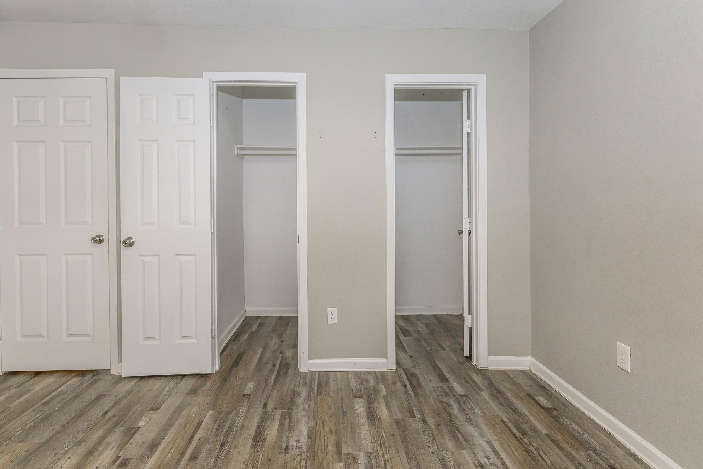 Interior view of a simple, empty room featuring light gray walls and a wood laminate floor. Two closets with white doors are on the left side, and one door leads to a separate space. Natural light brightens the area, creating a clean and spacious feel.
