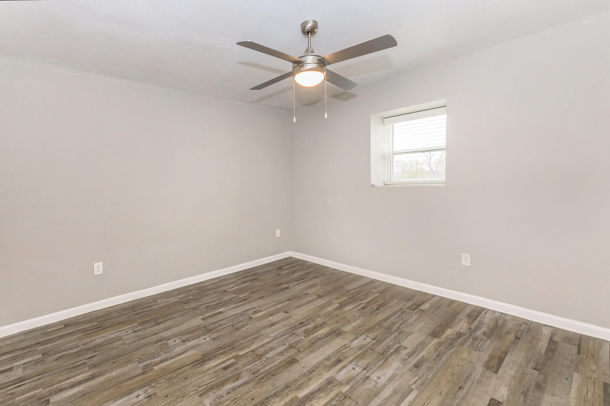 Empty room with light gray walls and a medium-toned wooden floor. A ceiling fan with a light fixture hangs in the center. A small window on the right allows natural light to enter. The space is uncluttered and ready for furnishing.
