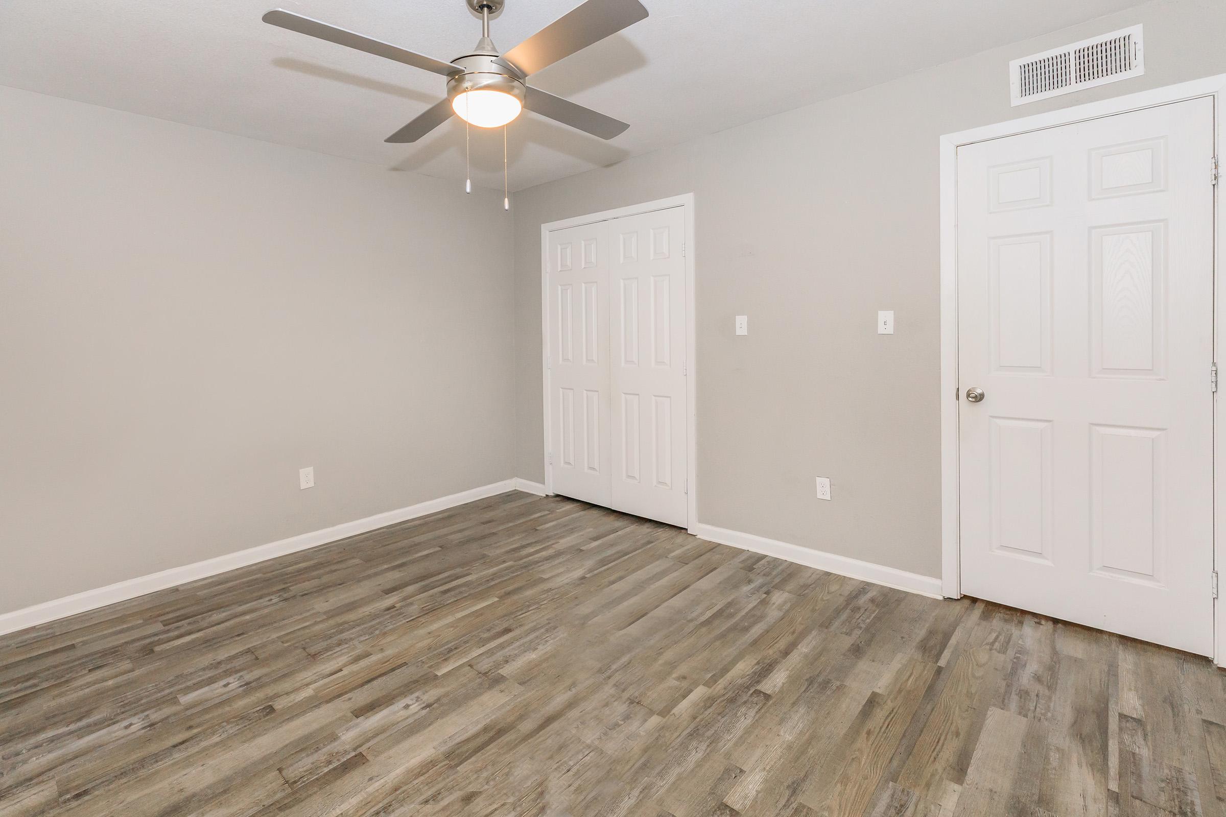 Empty room with light gray walls and wooden-looking laminate flooring. A ceiling fan is installed, and there are two closed white doors on one wall. The room is well-lit, giving it a spacious and clean appearance.