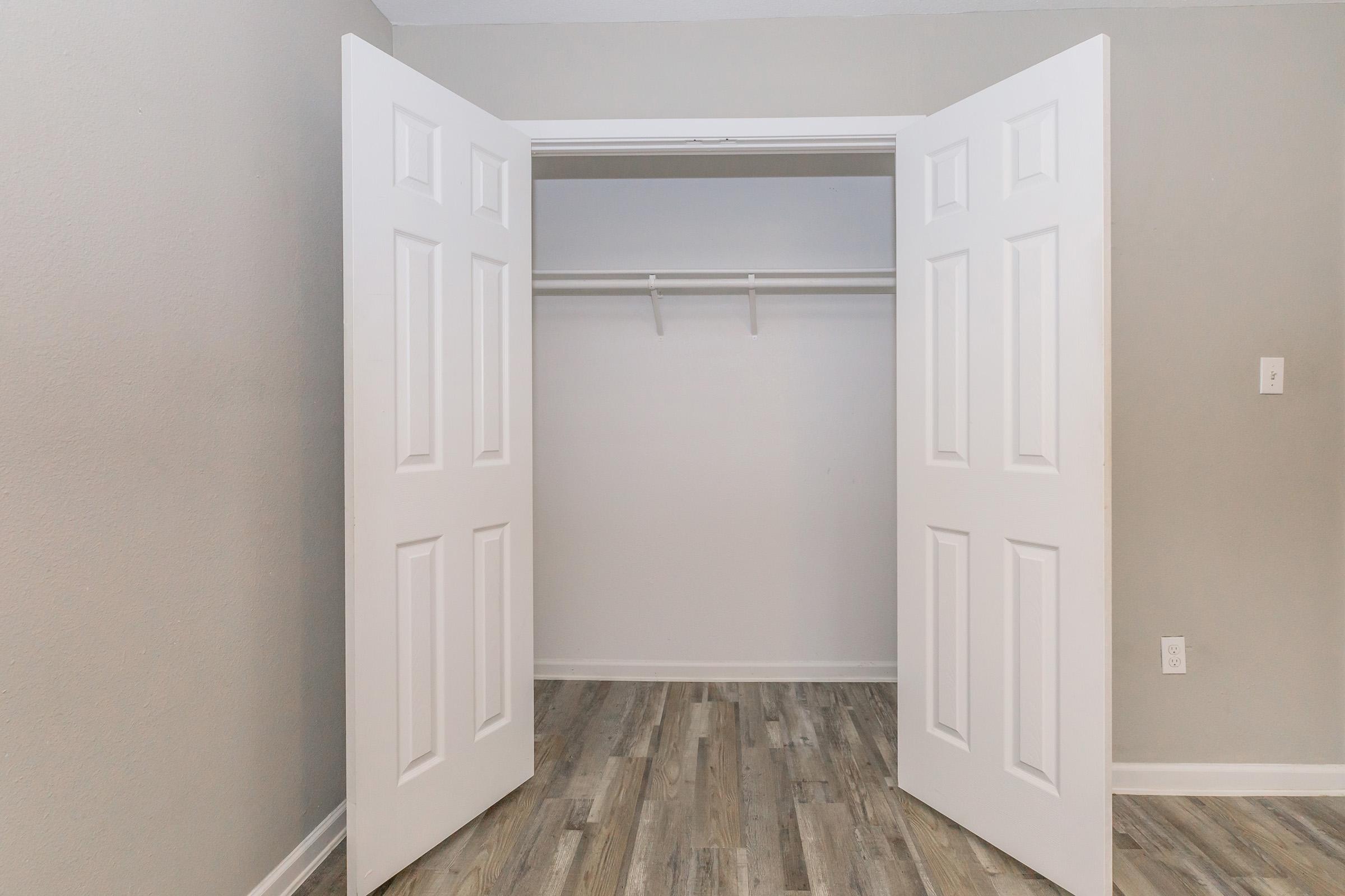 An empty closet with two open white doors against a light gray wall. The floor features wooden laminate in a natural tone. There is a single hanging rod inside the closet, indicating space for storage.