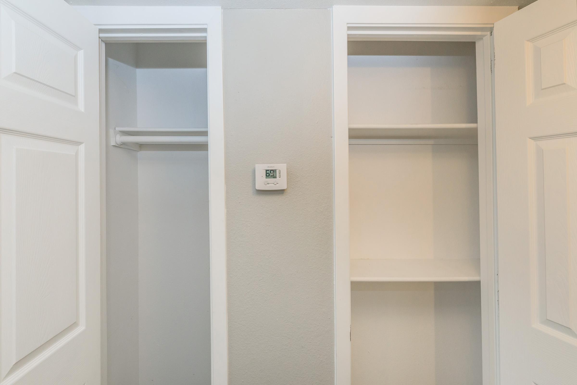 A photo of an empty closet space with two open doors, revealing built-in shelves on either side. A thermostat is mounted on the wall between the closets. The walls are painted in a light color, creating a clean and minimalistic appearance.