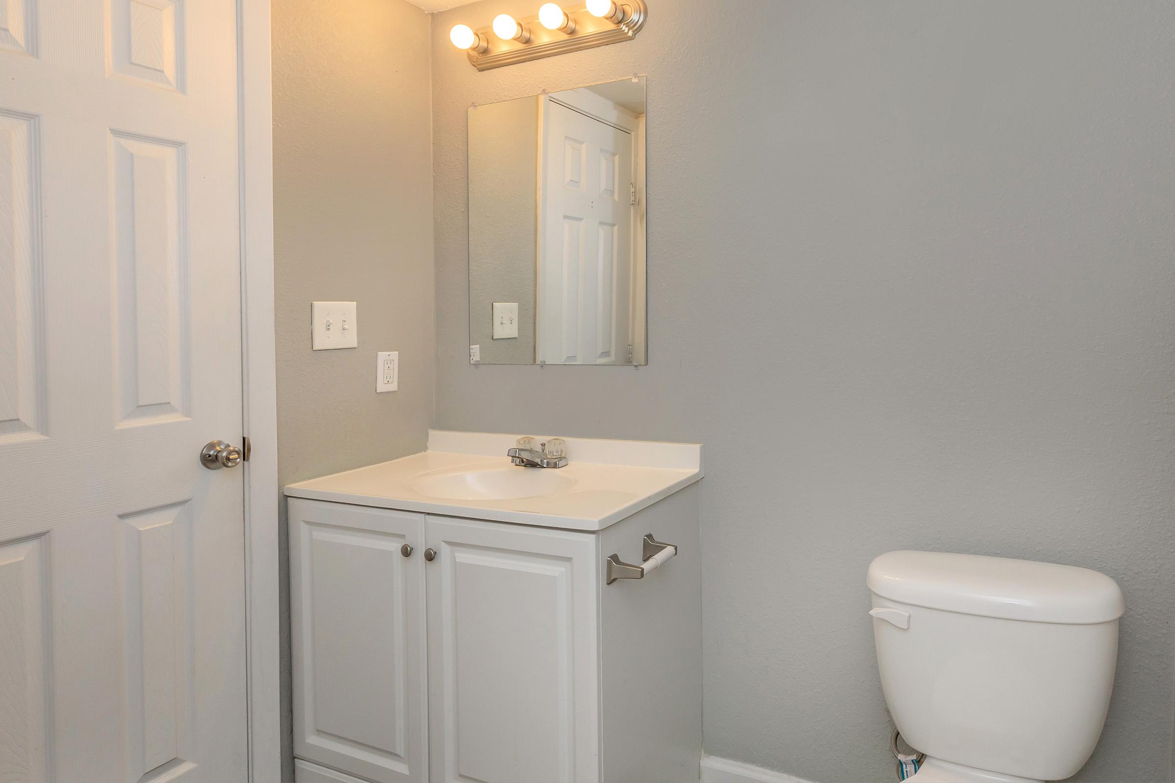 A small bathroom featuring a light gray wall, a white vanity with a sink, a large mirror above the sink, a white toilet, and a closed door. The lighting is provided by five bulbs above the mirror, creating a bright atmosphere. The overall design is clean and modern.