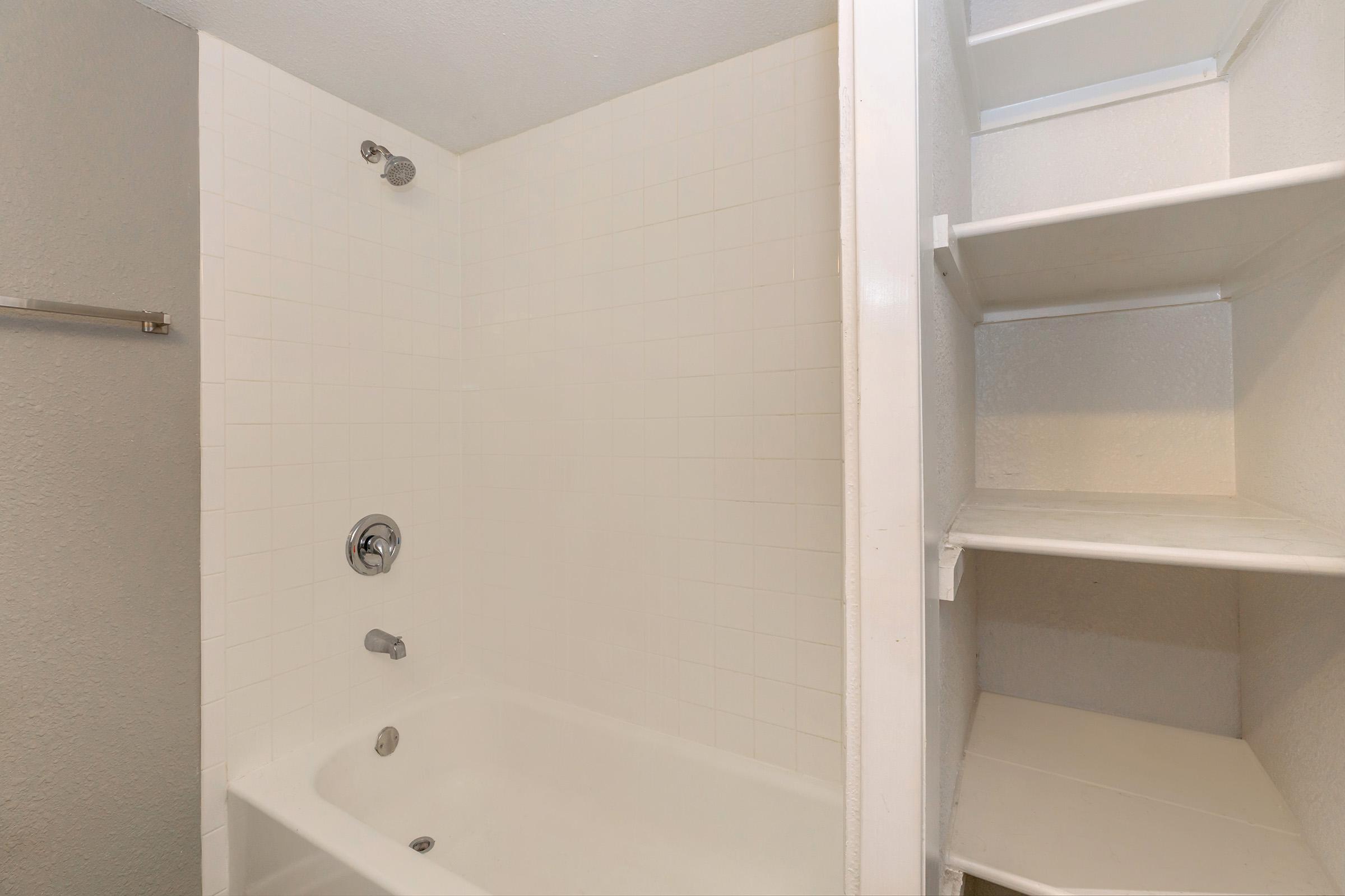 A clean, modern bathroom featuring a bathtub with a showerhead, white tiled walls, and a shelf unit for storage on the right. The overall aesthetic is simple and uncluttered, emphasizing a light and airy feel.