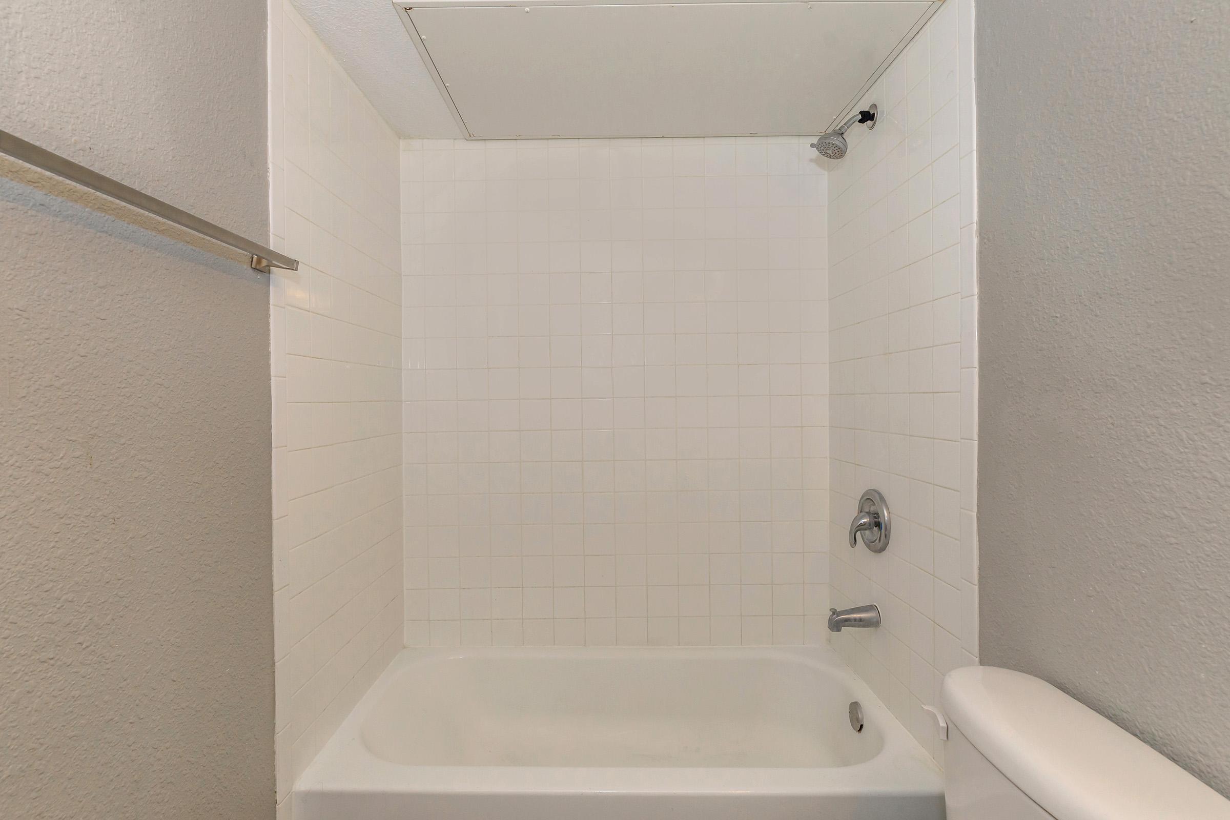 A clean and simple bathroom interior featuring a white tiled shower area with a bathtub, a single faucet, and a towel bar on the left side. The walls are painted in a light gray color, and the ceiling is plain and unobtrusive. Natural light may enter from the top, enhancing the overall brightness.