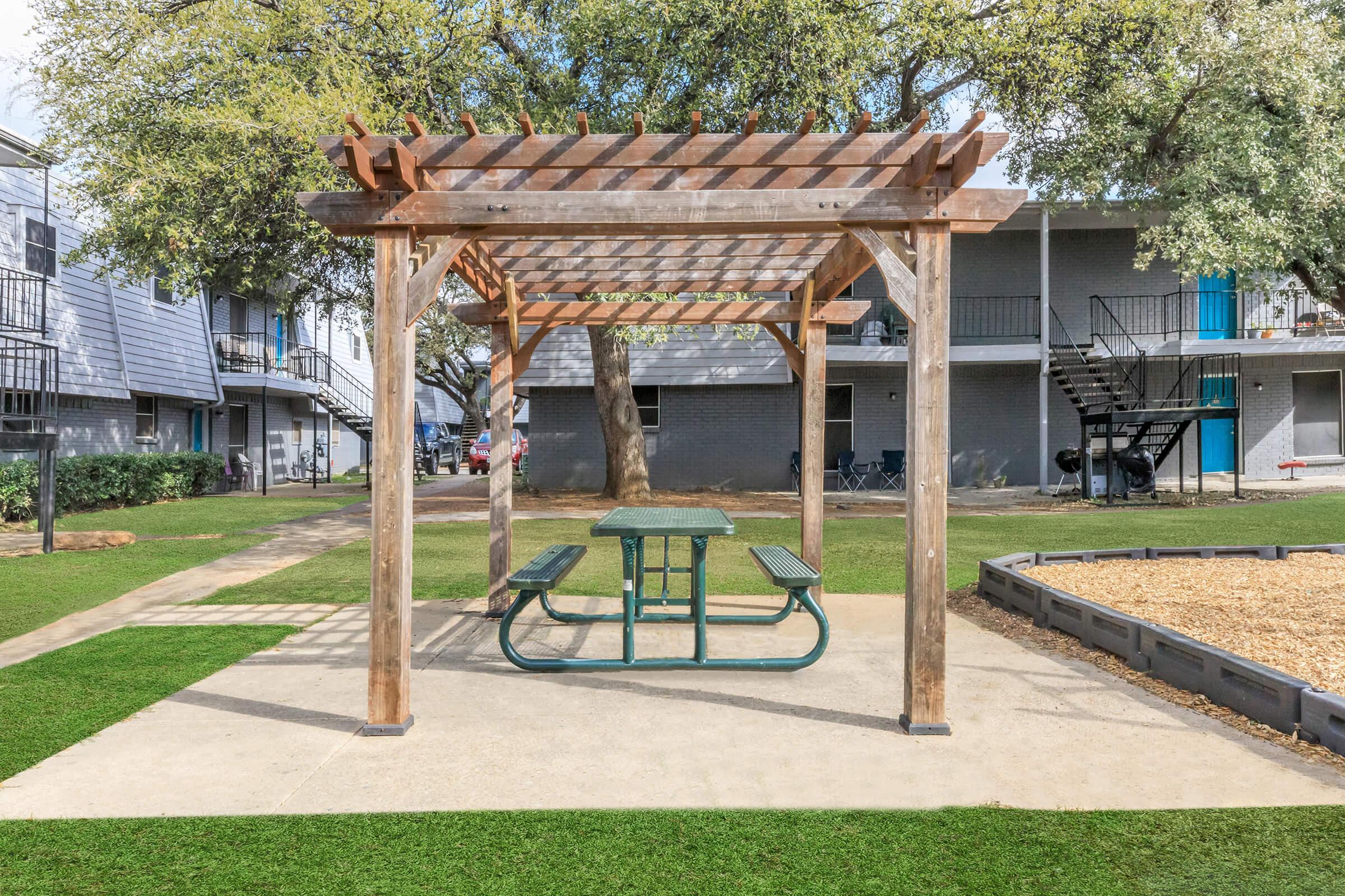 A wooden pergola with a green picnic table underneath is situated on a lawn in a residential area. In the background, there are gray buildings with balconies and a spiral staircase. Trees provide shade, and a gravel area is visible nearby.