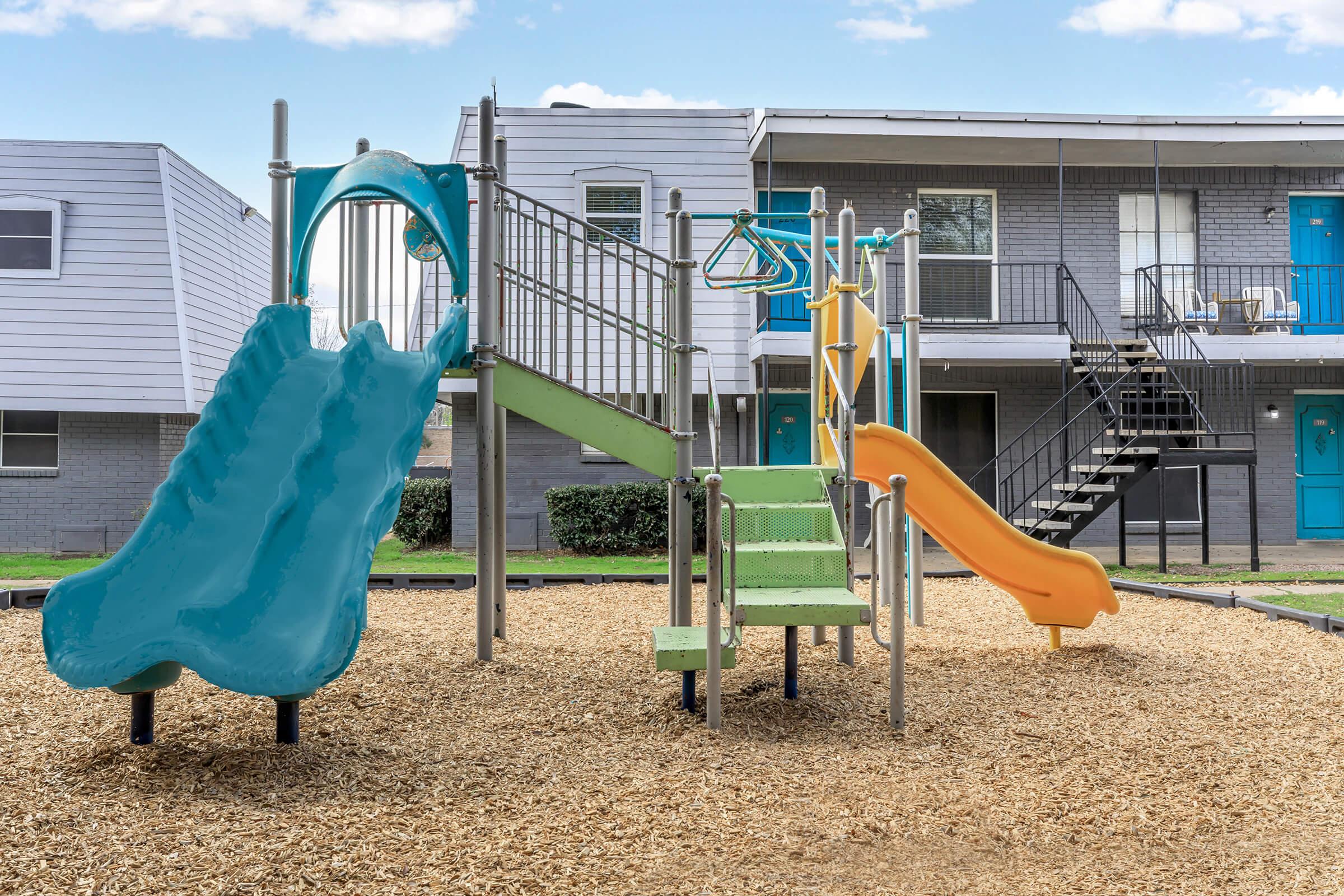 A playground featuring two slides in blue and orange, a green climbing structure, and monkey bars, surrounded by mulch and apartment buildings in the background. The scene is bright and inviting, perfect for children's outdoor play.