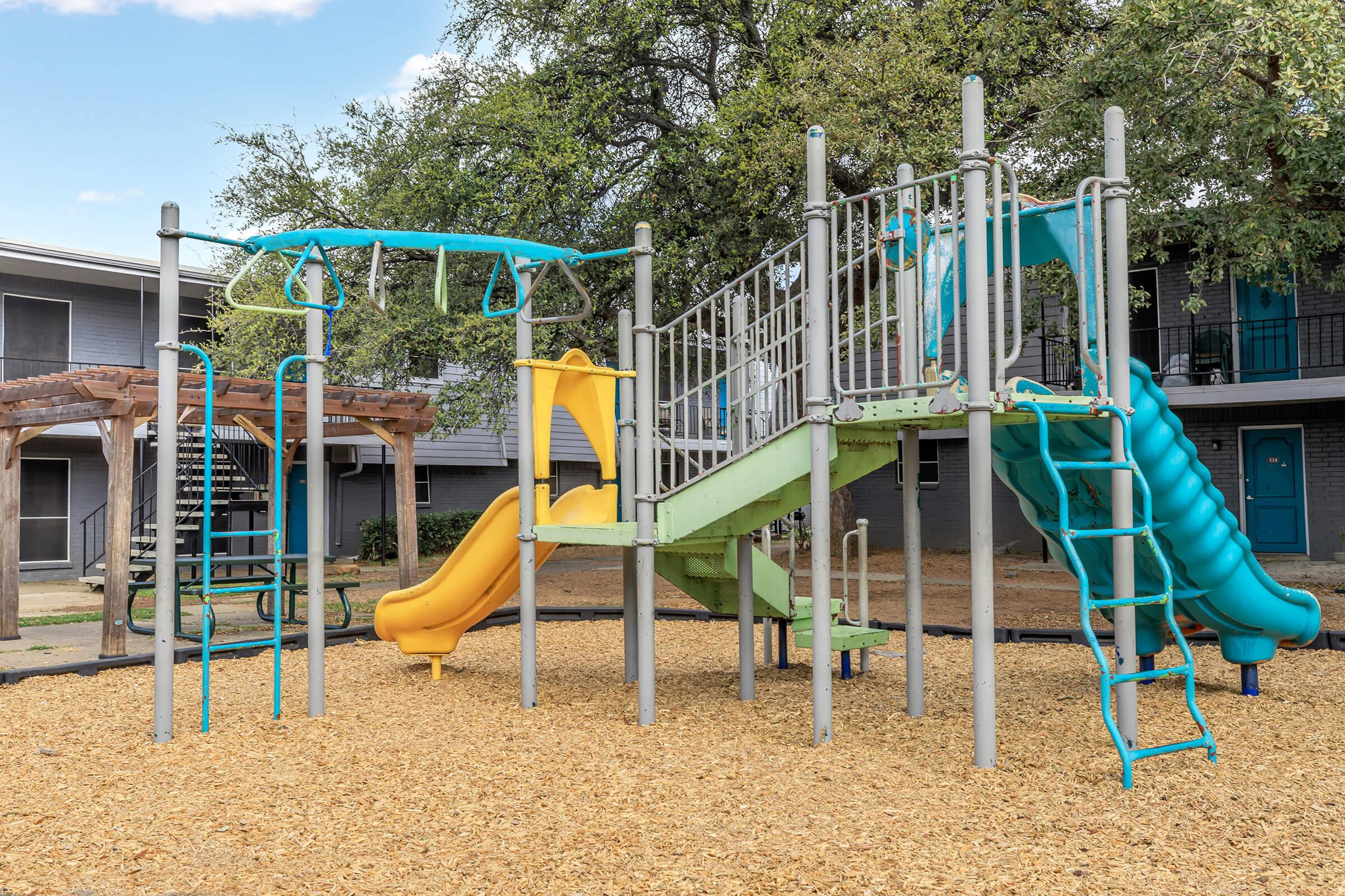A colorful children's playground featuring a green and yellow slide, climbing structures, monkey bars, and swings, surrounded by a sandy area. The playground is set in a community space with nearby benches and buildings in the background, providing a fun and safe environment for kids to play.
