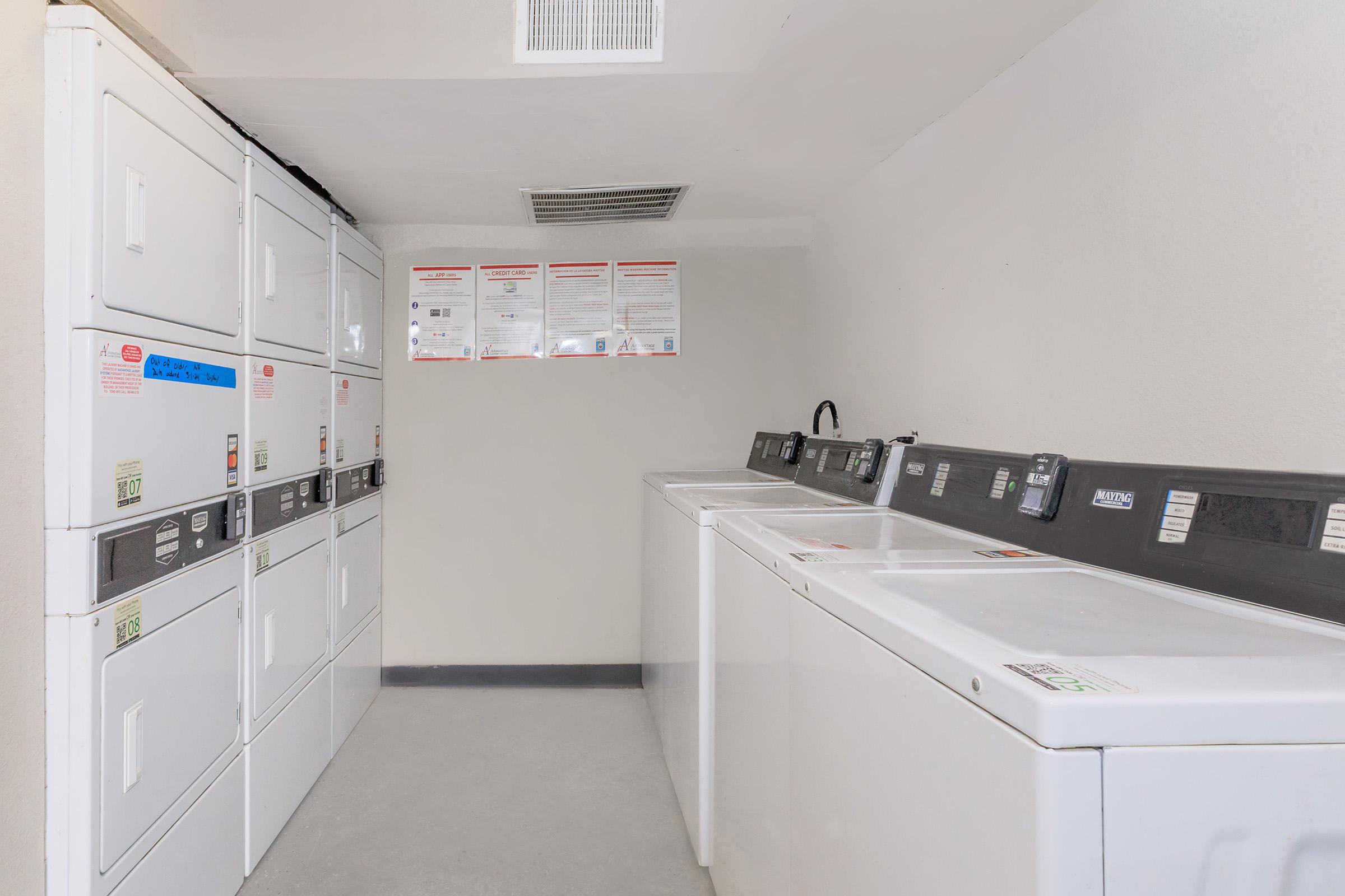 A clean and organized laundry room featuring several stacked washing machines and dryers against a light-colored wall. Instructions and safety notices are posted above the machines. The floor is gray, and the overall space is well-lit and tidy.