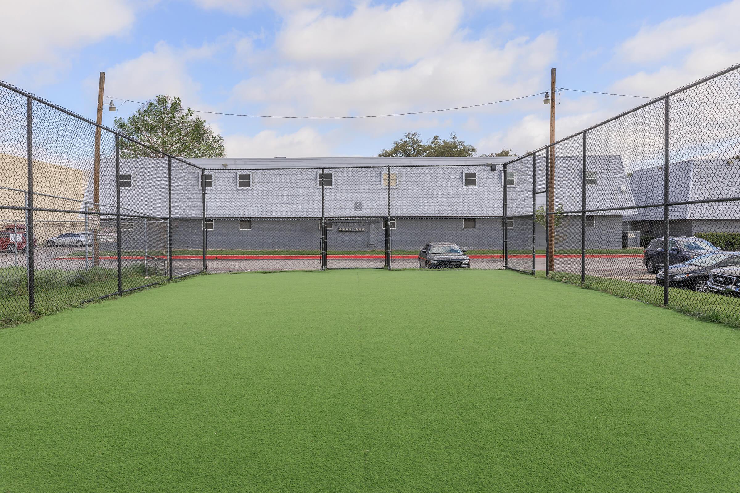 A fenced sports field with artificial turf, featuring a bright green surface. In the background, there are several buildings and a parked black car. The sky is partly cloudy, with some blue visible. The scene is set in an urban environment.